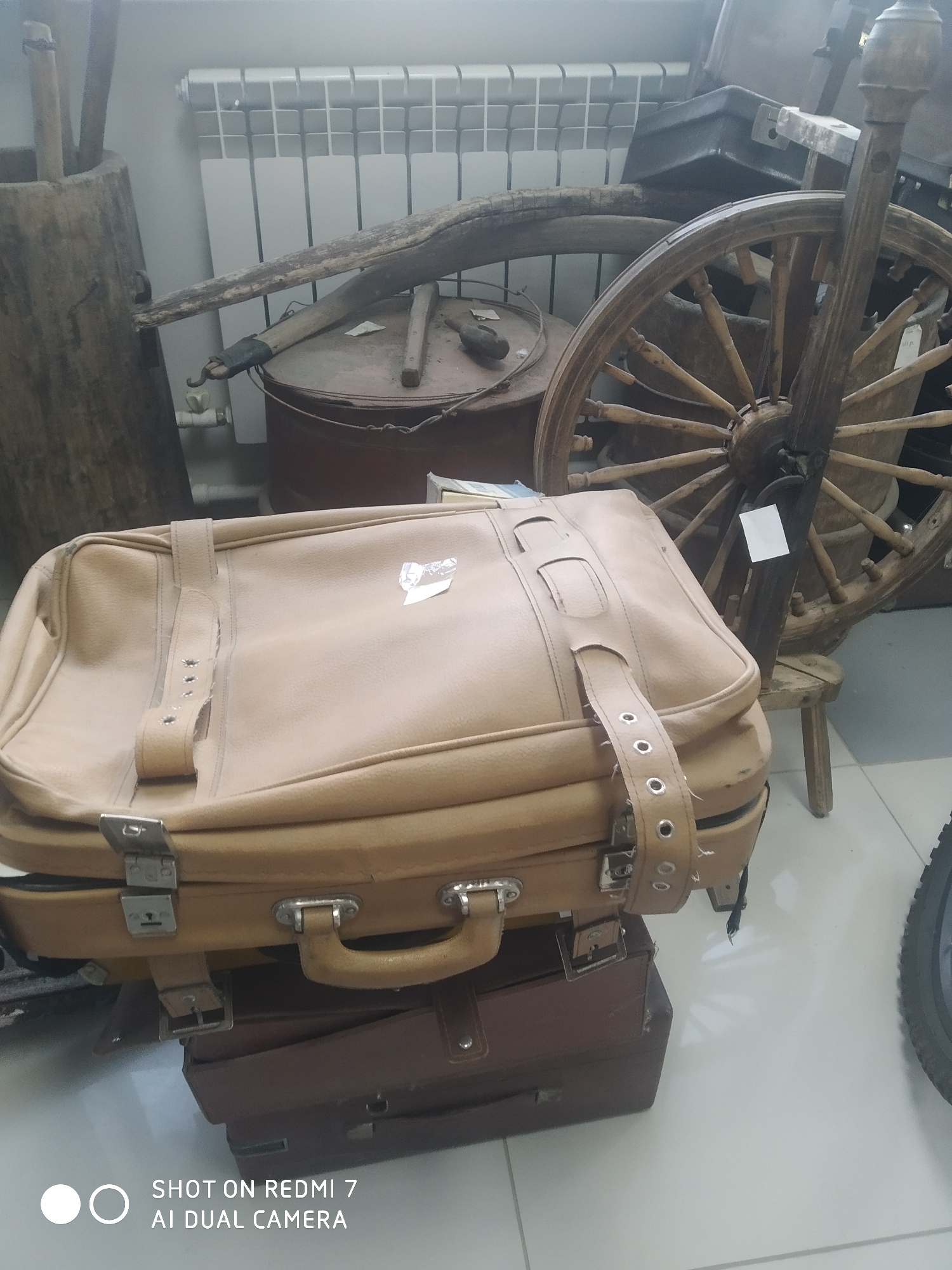 The image shows a stack of three vintage suitcases in what appears to be a museum or storage area in Chaikovsky, Russia. The top suitcase is light brown with straps and metal buckles, the middle one is a similar shade but darker, and the bottom suitcase is dark brown. Behind the suitcases are various old agricultural tools and equipment, including a wooden wheel, a wooden container, and a rusty metal container. A white radiator is visible on the wall. The floor is light-colored tile. The inscription "SHOT ON REDMI 7 AI DUAL CAMERA" is visible in the bottom left corner.