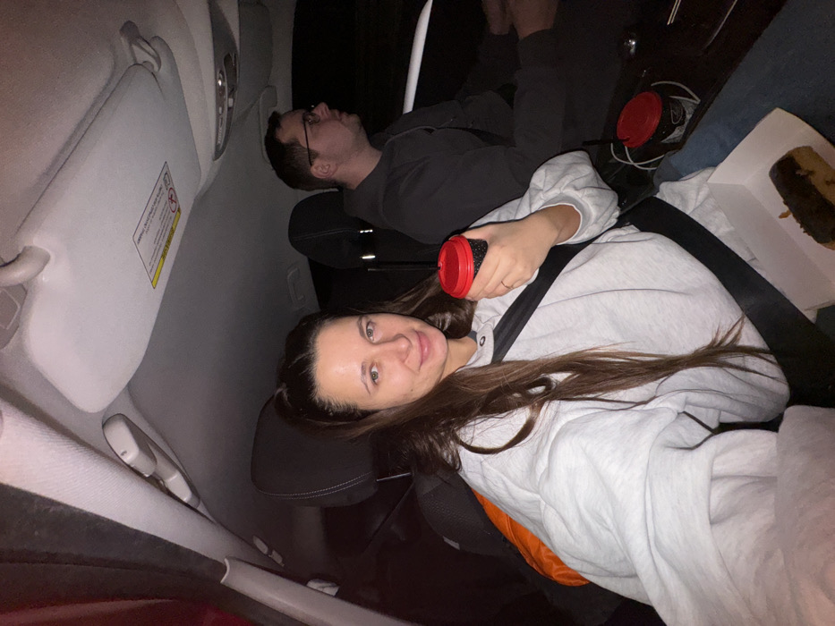 Inside a vehicle, a woman in a grey sweatshirt smiles at the camera, holding a red-lidded cup. She is wearing a seatbelt. Behind her, a man in a grey shirt reclines. A second red-lidded cup and a partially eaten dessert are visible. The car's interior includes a sun visor. The scene appears to be lit by artificial light.