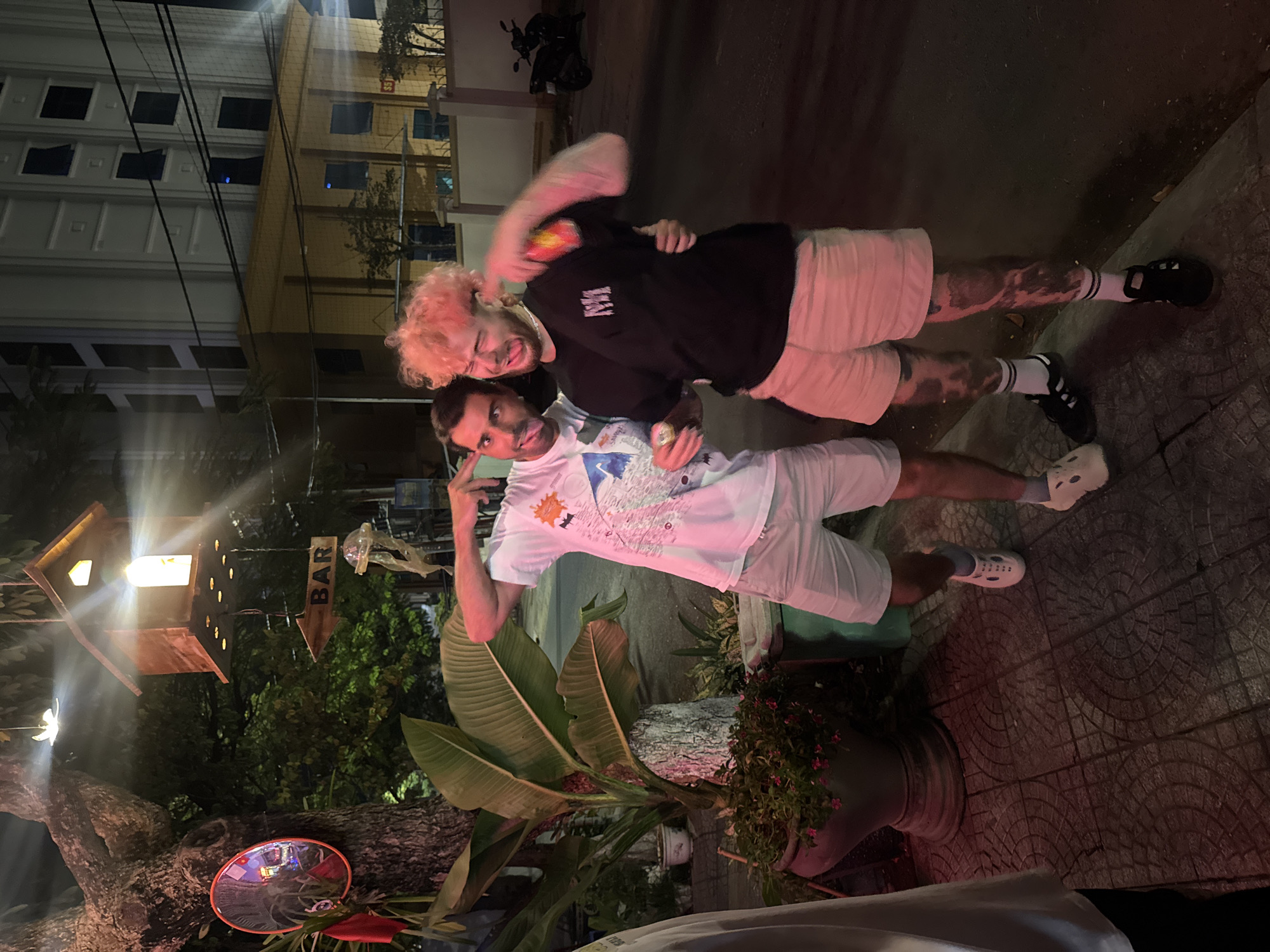 This vertical night-time photograph, taken in Đà Nẵng, Vietnam, captures two young men posing cheerfully on a patterned urban sidewalk. The scene is illuminated by artificial lights, casting warm glows on the subjects and surroundings.
The man on the left has distinctive light, possibly pinkish or blonde, curly hair. He wears a black t-shirt and light-colored shorts, with visible tattoos on his legs. He is embracing the other man, smiling and making a playful expression, while holding a small object, possibly a drink can. He wears dark athletic shoes with white socks.
The man on the right has dark hair and wears a white t-shirt with a colorful, abstract design, paired with light-colored shorts. He is also smiling and making a gesture with his right hand, holding a small, metallic can. His legs also feature tattoos, and he wears white, Croc-style sandals with light blue socks.
In the background, multi-story buildings with windows are visible, along with overhead utility wires. A motorbike is parked further down the street. To the left, a large tree trunk supports a round, convex mirror and lush green foliage. A prominent wooden sign, shaped like an arrow and featuring a small house-like structure with a light, clearly displays the word 'BAR'. Various potted plants with large leaves and small pink flowers decorate the scene, suggesting a vibrant, possibly entertainment-focused, area. The men appear to be friends enjoying a casual evening out, displaying a playful and cheerful mood.