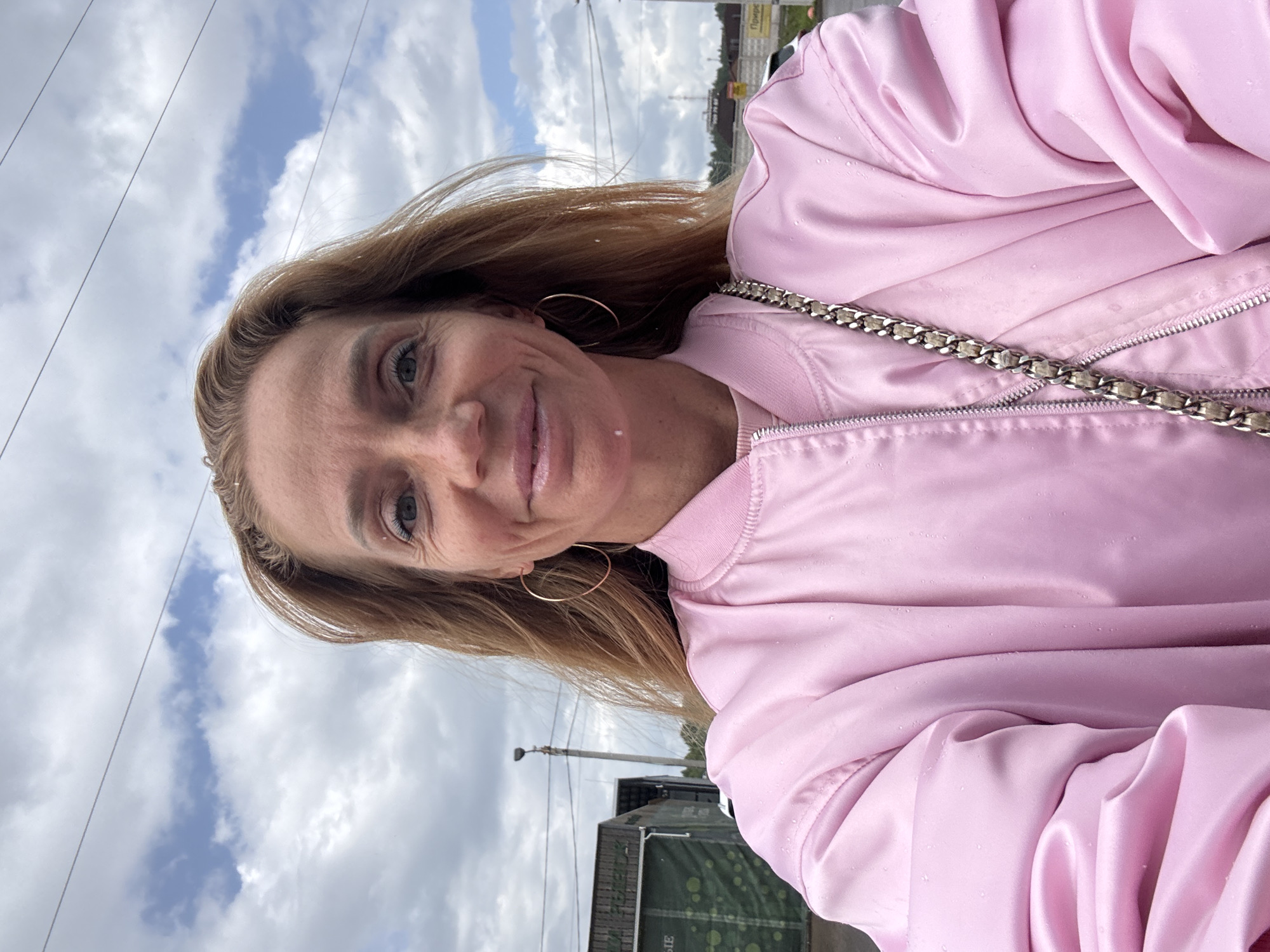 This is a selfie of a woman with blonde hair, wearing a pink satin bomber jacket. She has a light smile and is looking towards the camera. Her left eye is visible, and she is wearing a hoop earring. The jacket has a silver chain detail over the zipper.
The background suggests an outdoor setting with a cloudy sky and power lines. A lamppost and some buildings are visible in the distance, one of which has a green banner with white text. The overall impression is of a casual, possibly impromptu, outdoor photograph. The weather appears overcast, with some visible droplets on the jacket suggesting it might be damp, possibly from rain or humidity. The time of day is not definitively clear, but the lighting suggests daytime. The provided location context is Medovoye, Russia.