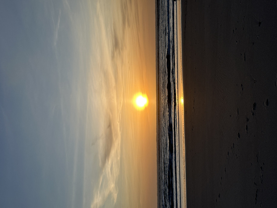 The image displays a sunset over a coastal beach in Gafanha da Nazaré, Portugal. The sky, occupying the upper two-thirds of the frame, exhibits a gradient from pale blue at the zenith to vibrant orange and yellow at the horizon, interspersed with faint, wispy clouds. The sun, a brilliant yellow-orange sphere, is positioned centrally, just above the ocean horizon. The dark blue-grey ocean shows moderate wave activity with distinct breaking waves near the shoreline. A luminous, elongated reflection of the sun stretches across the water towards the foreground. The lower third of the image features dark, damp sand, clearly imprinted with multiple human footprints. No human figures or specific objects are visible within the scene.