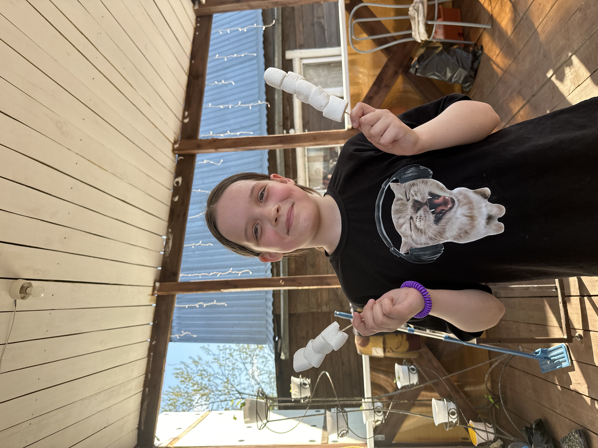 A young person is holding two sticks with marshmallows on them, indoors on a wooden porch-like area. The person has fair skin, light brown hair pulled back, and is wearing a black t-shirt with a cat wearing headphones on it, and a purple bracelet. The sticks have about four marshmallows each. The background shows a wooden structure, possibly an overhang or porch, with string lights and a corrugated metal roof. There's a glimpse of the outside with foliage and a white building. The scene is oriented vertically. The city Chaikovsky, Russia is not identifiable from this image.