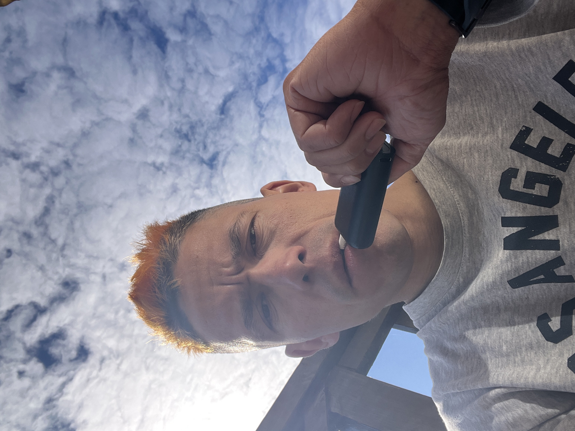 A low-angle shot depicts a person holding a vaping device to their lips, against a backdrop of a partly cloudy sky. The person has short, dyed orange hair. They are wearing a gray t-shirt with the word "LOS ANGELES" printed on it in black, visible partially. The location is not apparent in the image; Chaikovsky, Russia is not identifiable based on the image content.