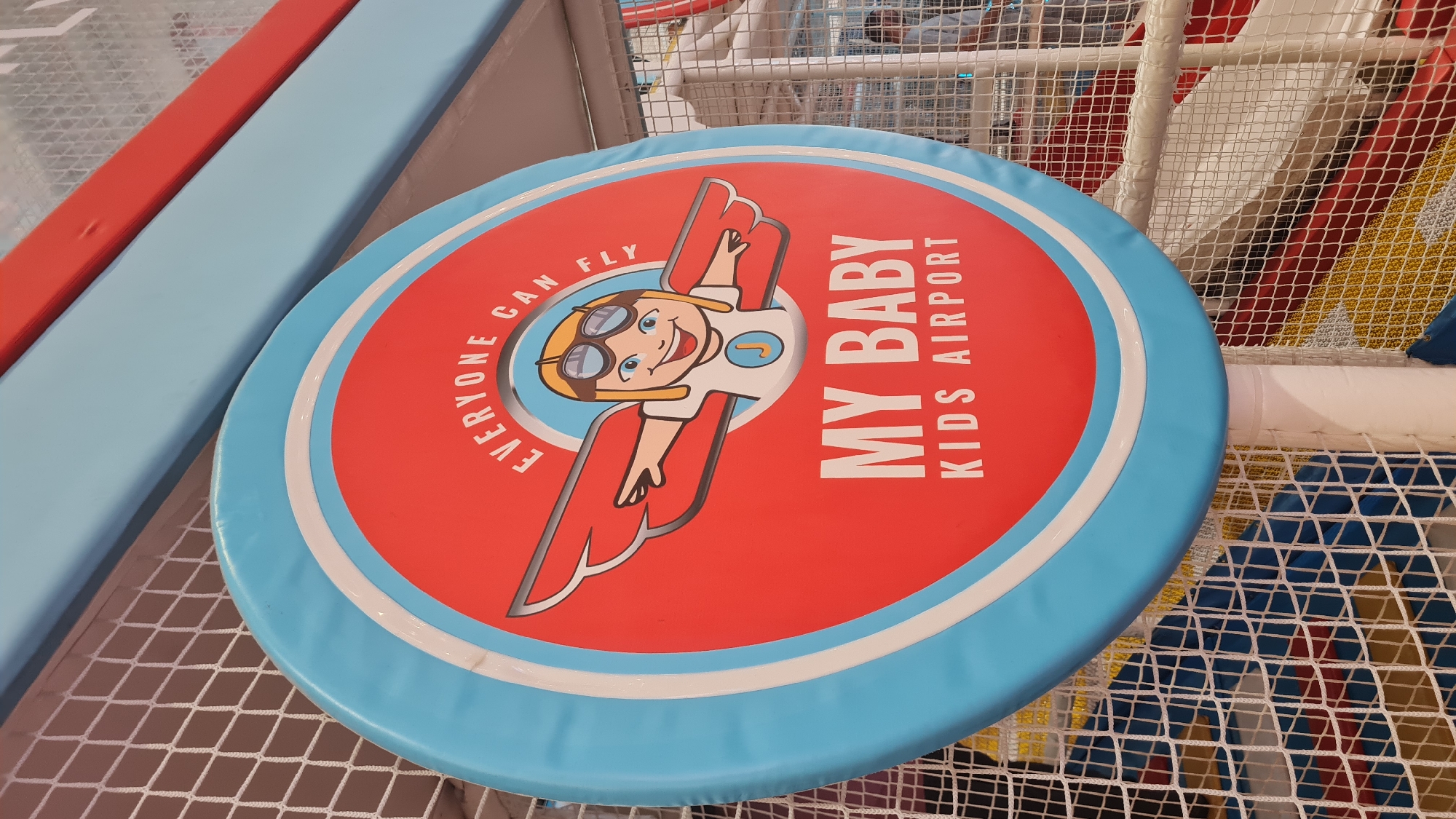 The image captures a close-up of a circular sign within an indoor children's play area, likely a soft play gym. The sign, predominantly red with a light blue and white border, features a cartoon character: a smiling child wearing pilot goggles and headgear, depicted with airplane-like wings extending from their back. The child has blonde hair and blue eyes, and a light blue circle with a "J" on it is visible on their chest.
Text is prominently displayed on the sign. Vertically aligned on the right side of the red circle, it reads "MY BABY KIDS AIRPORT" in white capital letters. Curved along the top-left edge of the red circle, the phrase "EVERYONE CAN FLY" is written in white capital letters, following the curve of the sign.
The background reveals typical elements of a children's play structure. Padded surfaces in light blue and red are visible, likely forming walkways or barriers. White safety netting is stretched across parts of the background, indicating an enclosed and safe play environment. The netting suggests multiple levels or sections within the play area. The lighting appears to be indoor and even, with no discernible time of day or weather conditions. The overall impression is that of a bright, cheerful, and engaging space designed for children's play, emphasizing themes of flight and adventure. The location is Yirka, Israel.