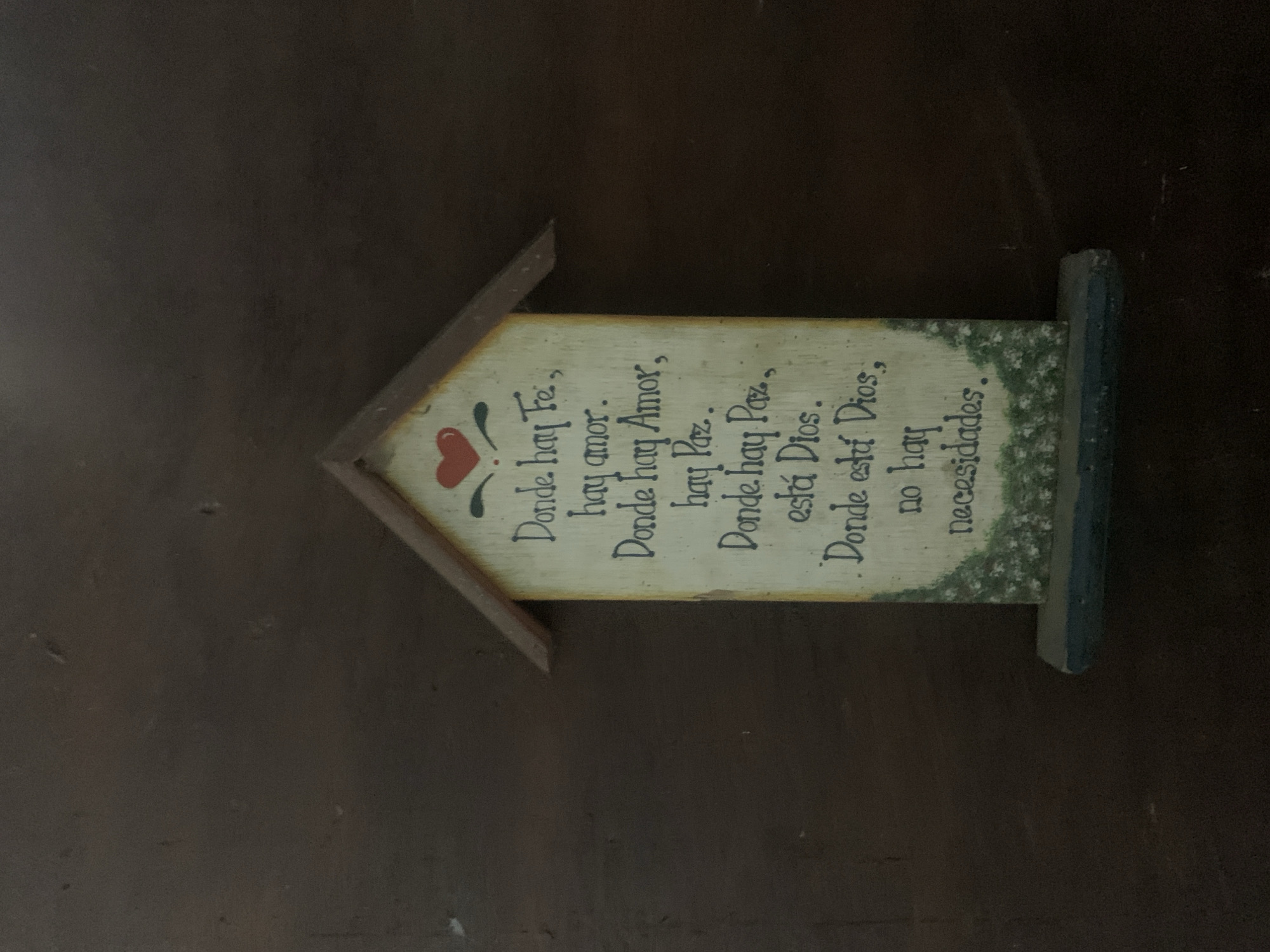 The image features a decorative wooden plaque, oriented horizontally on a dark, indistinct background. The plaque is shaped like a house, with a cream-colored, rustic-textured main body. It has a pointed, reddish-brown roof section on the left and a blue-painted base on the right. A small red heart, adorned with green and black vine-like flourishes, is painted near the top left edge of the plaque's surface.
The plaque is inscribed with an inspirational quote in Spanish, handwritten in a decorative black script. The text reads: "Donde hay Fe, / hay amor. / Donde hay Amor, / hay Paz.. / Donde hay Paz, / está Dios. / Donde está Dios, / no hay / necesidades." This translates to English as: "Where there is Faith, / there is love. / Where there is Love, / there is Peace. / Where there is Peace, / there is God. / Where there is God, / there are no / needs." The background appears to be a dark surface, possibly a table or shelf, in an indoor setting with subdued lighting. This item is located in Santa Ana, Venezuela.