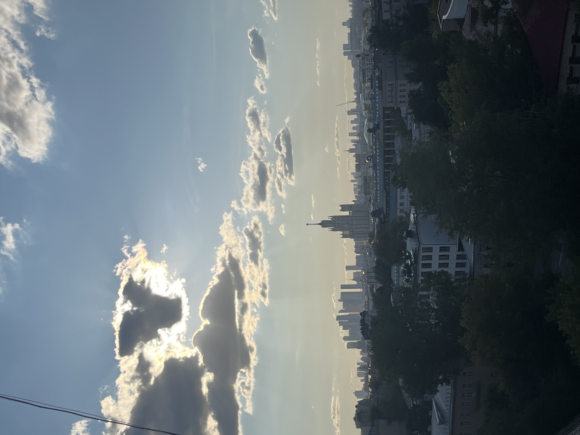This is a medium-shot, outdoor, daylight photograph of a city skyline, likely Moscow, Russia. The sky is a bright blue with scattered white and gray clouds. The sun is partially obscured by a dark cloud in the upper left quadrant, casting rays of light downwards and creating a halo effect. In the distance, a cluster of modern skyscrapers dominates the horizon. To the right of the skyscrapers, a tall, ornate building with a spire, resembling a Stalinist skyscraper, stands prominently. In the foreground, lush green trees and the roofs of various buildings are visible, suggesting a residential or mixed-use urban area. The overall lighting suggests either early morning or late afternoon. There are no people or animals visible in the image.