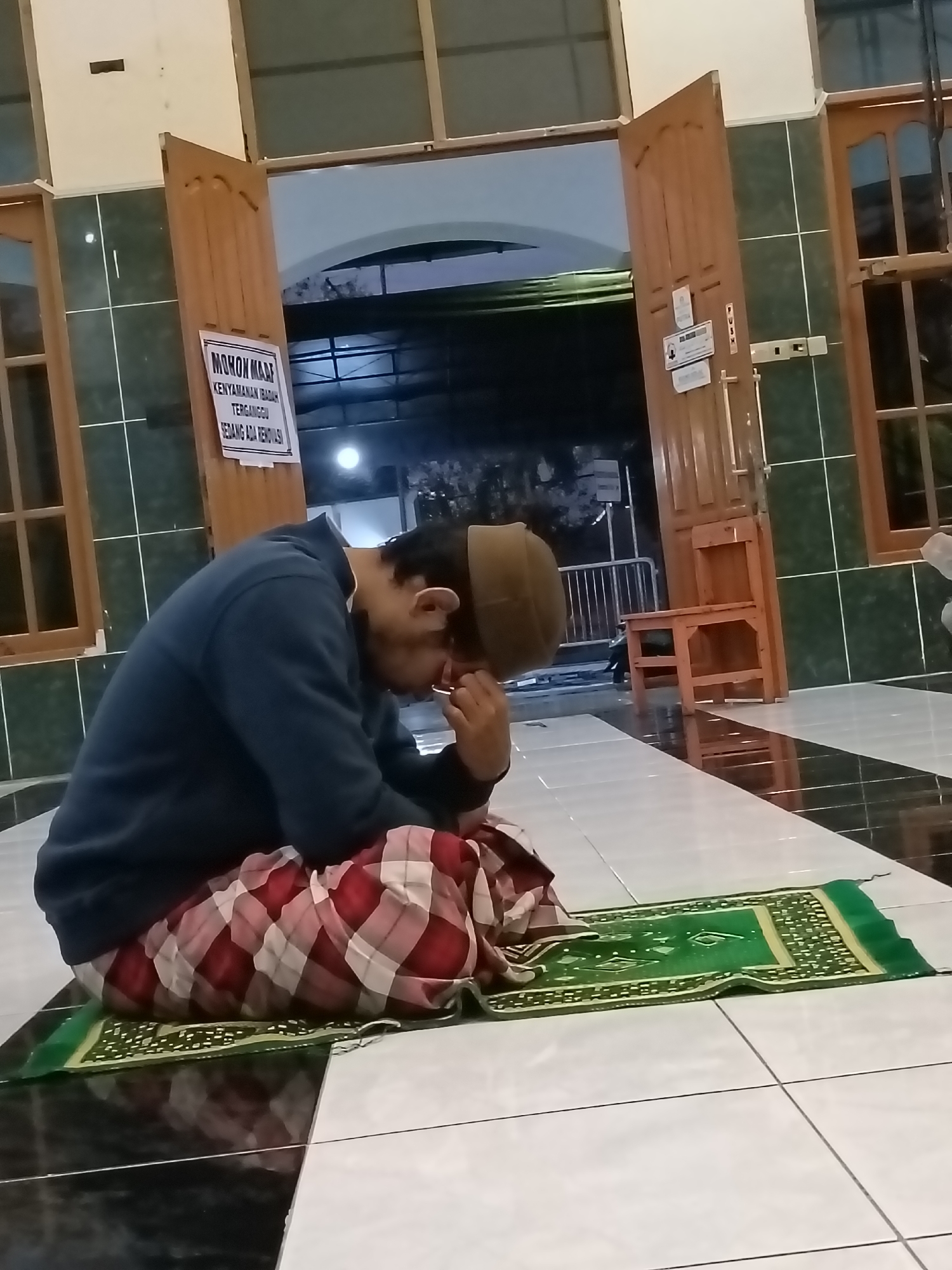In Kediri City, Indonesia, a person wearing a brown peci and a blue sweatshirt is sitting on a green prayer mat on a tiled floor. They are also wearing a checkered sarong in red and white. The person has their head bowed and appears to be covering their face with their hand. The background features wooden doors, a chair, and a sign that reads "MOHON MAAF KEAMANAN IBADAH TERGANGGU SEDANG ADA RENOVASI," which translates to "SORRY, PRAYER SECURITY IS DISTURBED, RENOVATION IS UNDERWAY." The lighting suggests it is nighttime or an indoor space with artificial lighting.