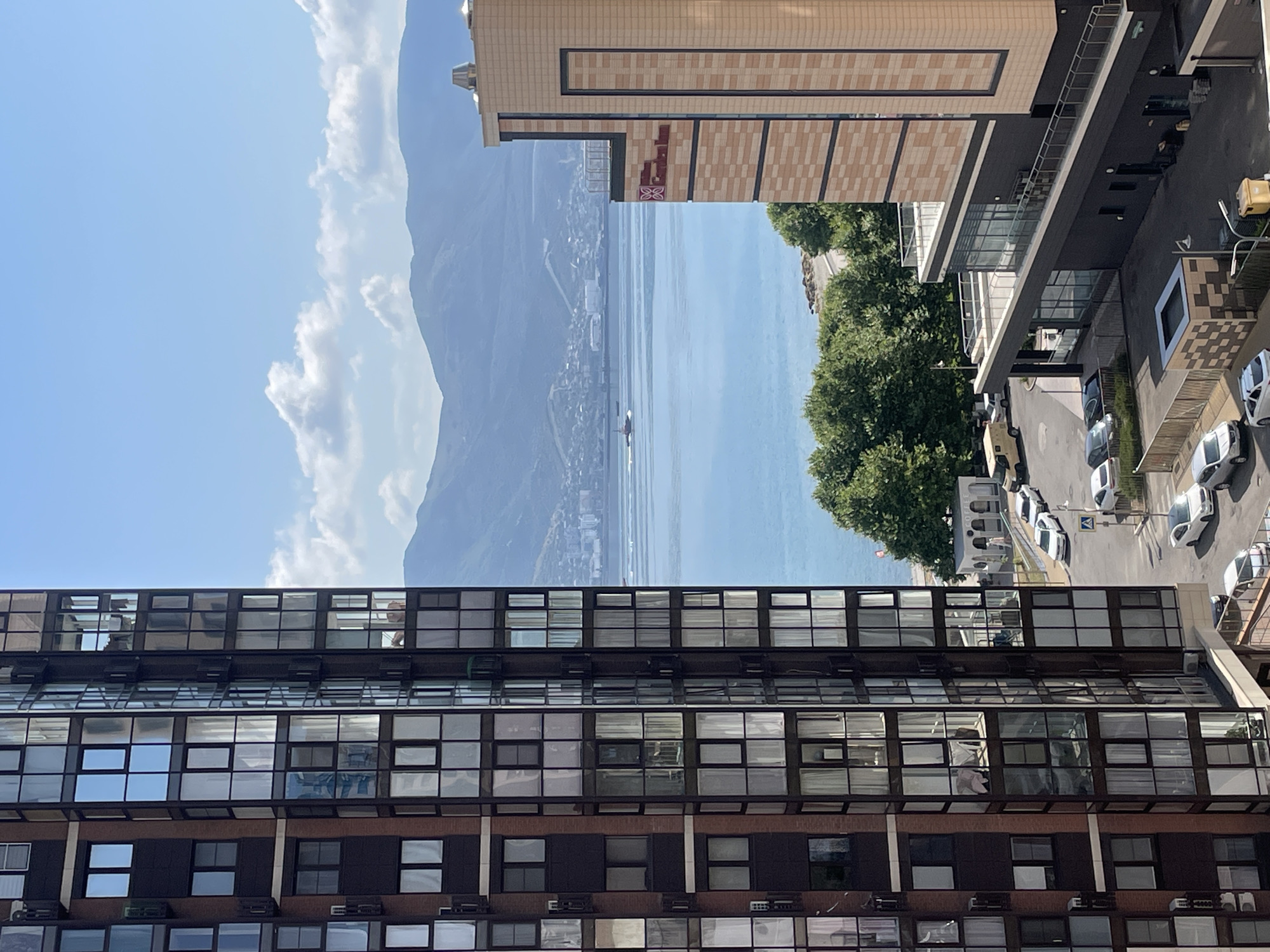 In an outdoor urban scene, a view encompasses a body of water with distant mountains under a blue sky with scattered clouds. In the foreground, a tall, modern building with a dark facade and numerous windows dominates the left side of the frame. To the right, a lighter-colored, multi-story building with a patterned facade and balconies is visible. Between the buildings and the water, a street with several parked cars and a yellow van is present, along with a line of lush green trees. A small boat is visible on the water in the middle ground.