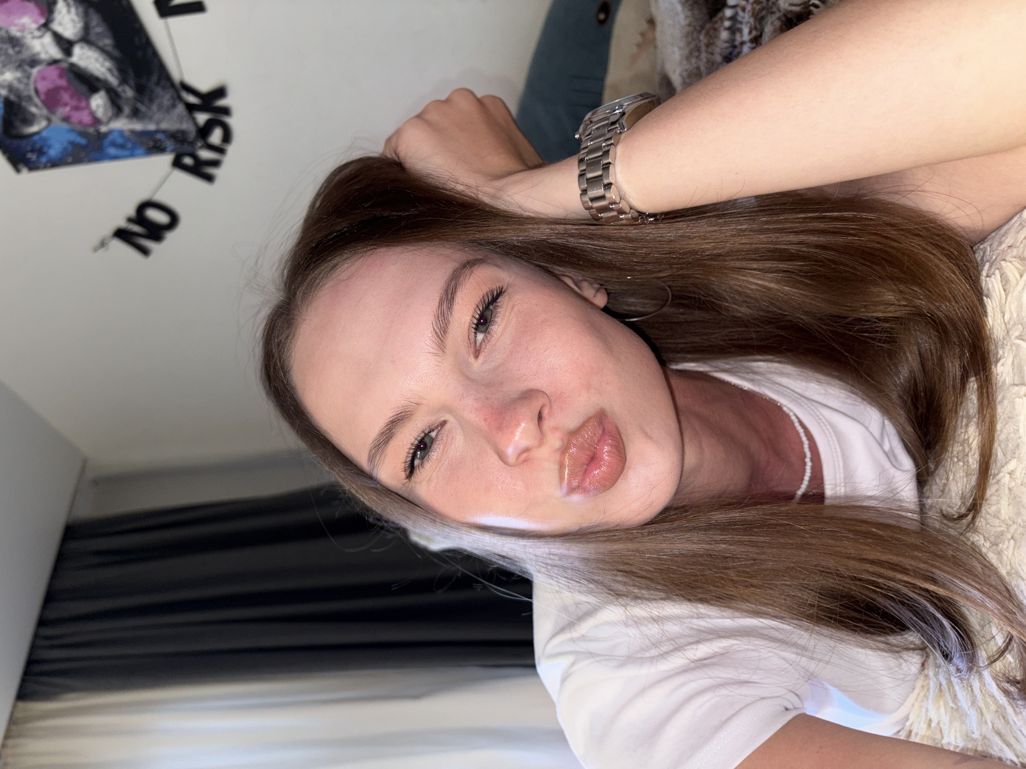 The image features a young adult female in an indoor setting, specifically a room within a residence in Perm, Russia. She has long, straight, brown hair, parted to the left, which cascades over her shoulders and chest. Her face exhibits a light complexion with apparent blush on her cheeks and nose. She is looking directly at the camera with light-colored eyes, displaying faint smile lines around them, and her lips are puckered in a "kissing" expression. She wears a plain white short-sleeved t-shirt, a small white beaded necklace, and a silver-toned hoop earring in her left ear. A large silver-toned watch with a metal link bracelet is visible on her right wrist, as her right hand is positioned behind her head, supporting it. She appears to be lying on a light-colored, fluffy textured blanket or rug. The background on the left side reveals a window covered by dual curtains, one light cream/white and the other dark gray/black. On the right side of the background, a white wall is adorned with a square piece of art depicting a cat's head with pink sunglasses and blue/purple accents. Below this artwork, a string of dark, cut-out letters spells "NO RISK". A patterned blanket and a blue cushion are partially visible behind her on the right.