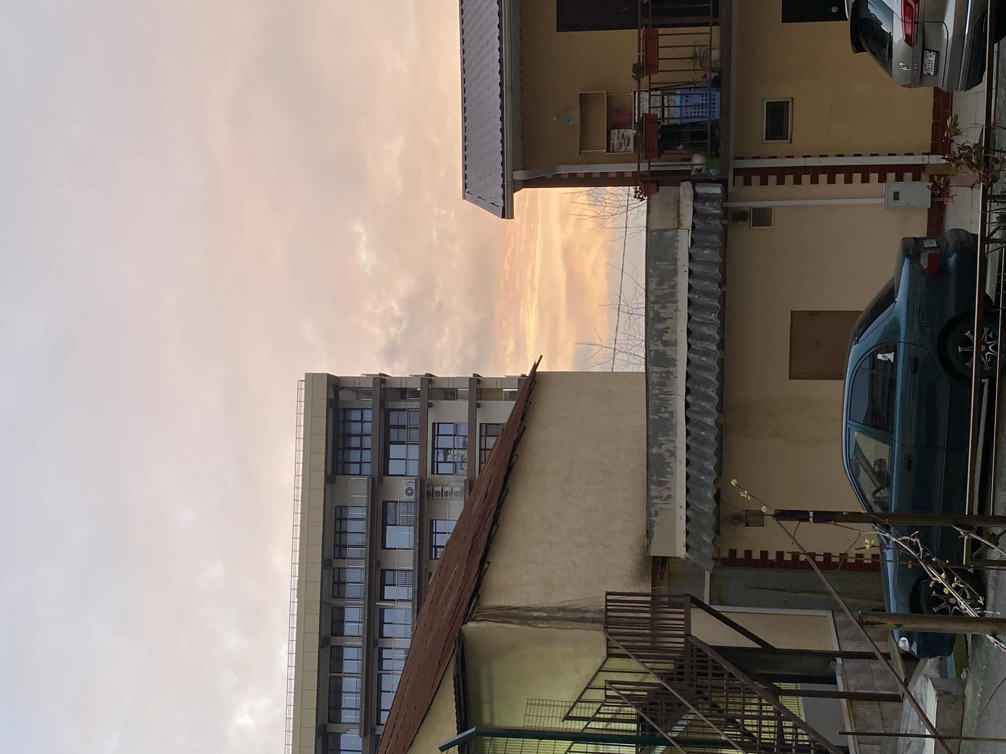 The image shows a building with a facade of windows and air conditioning units in Sochi, Russia. A narrow alleyway is visible with other buildings on either side. There are cars parked in the alley. The sky is cloudy with the sun breaking through. The buildings are varying heights and construction types. The focus is on the architecture and urban environment.