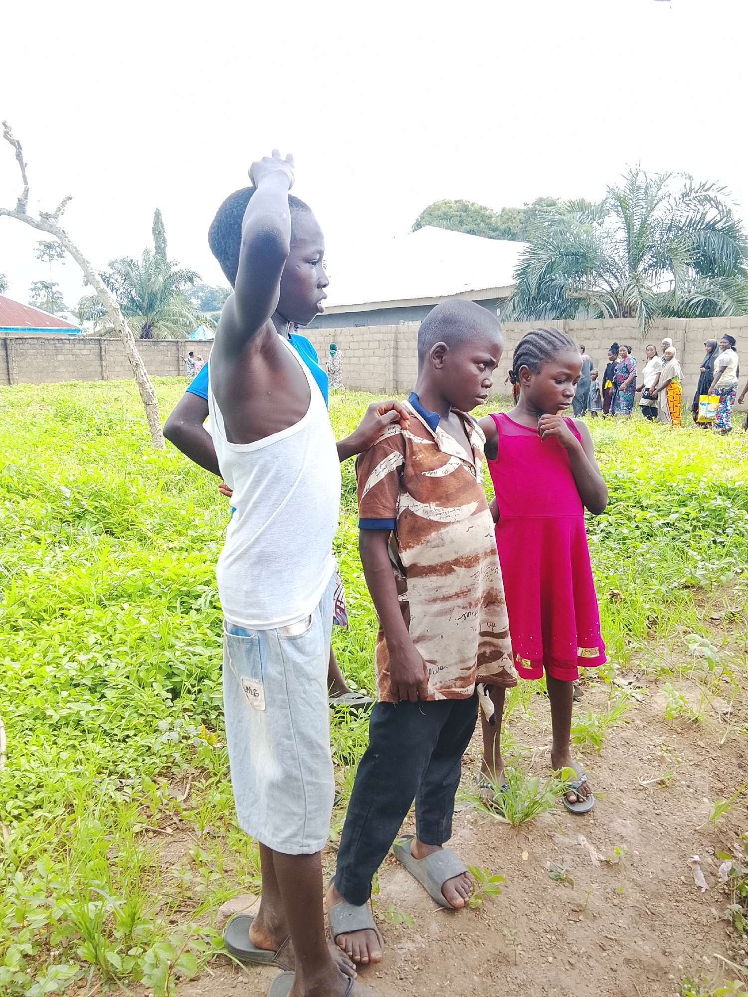 In Akwanga, Nigeria, three children stand on a dirt path surrounded by lush green vegetation and a brick wall. The child on the left, wearing a white tank top and denim shorts, is looking upwards and to the right with their left arm raised. To the right of them, a young boy in a brown and white patterned shirt and black pants stands with his right arm around the shoulders of a young girl in a bright pink dress. In the background, several adults are visible, some carrying items, gathered near a building. The sky is overcast, suggesting cloudy weather.