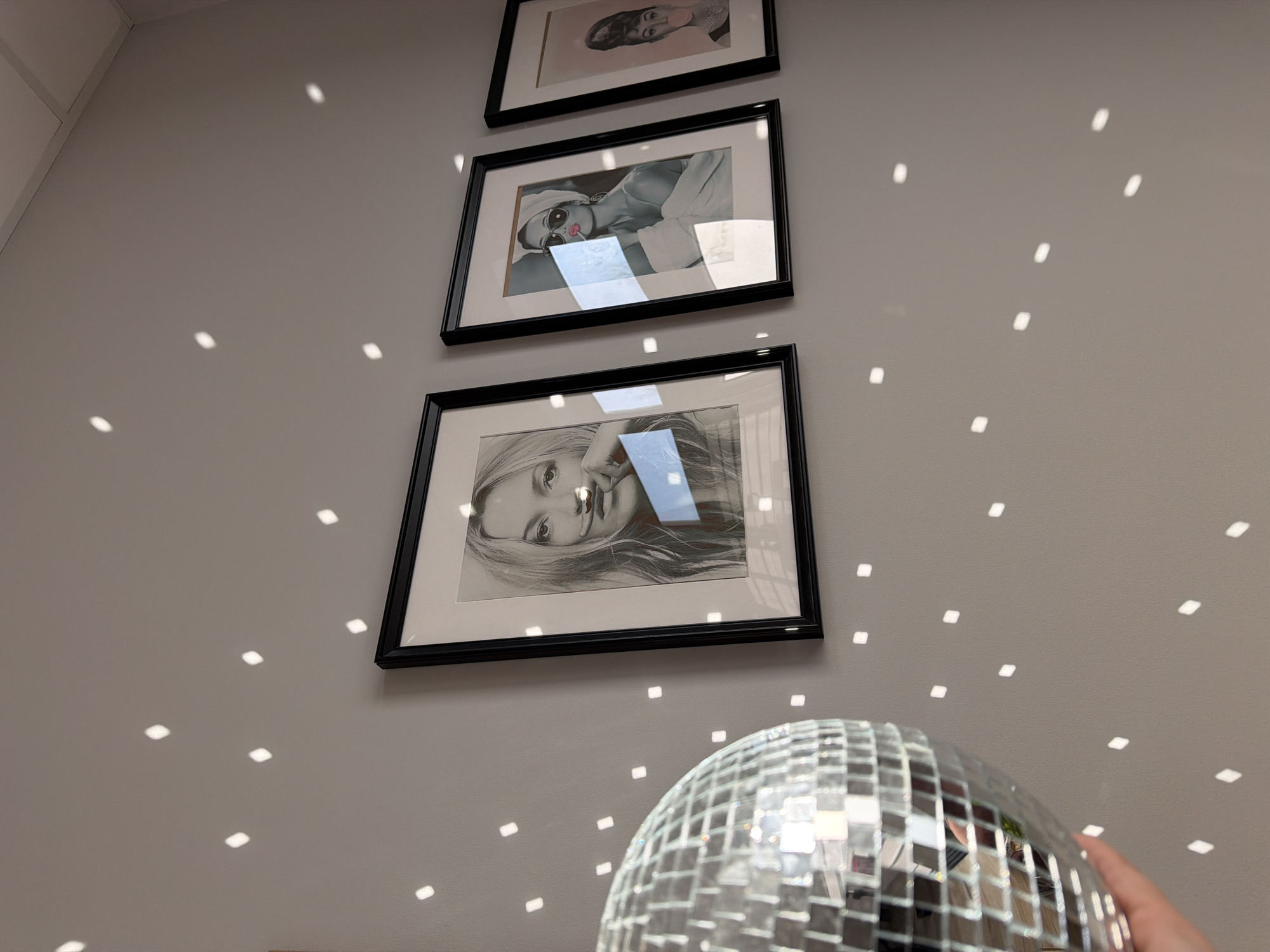 The image shows a section of a wall adorned with three framed pictures and a disco ball. The pictures, hanging vertically, feature portraits of women. The disco ball is partially visible in the lower portion of the image, reflecting light onto the surrounding wall, creating a pattern of small, square highlights. A hand is partially visible holding the disco ball. The wall is a light neutral color. There is no information to identify the city or country of the location.