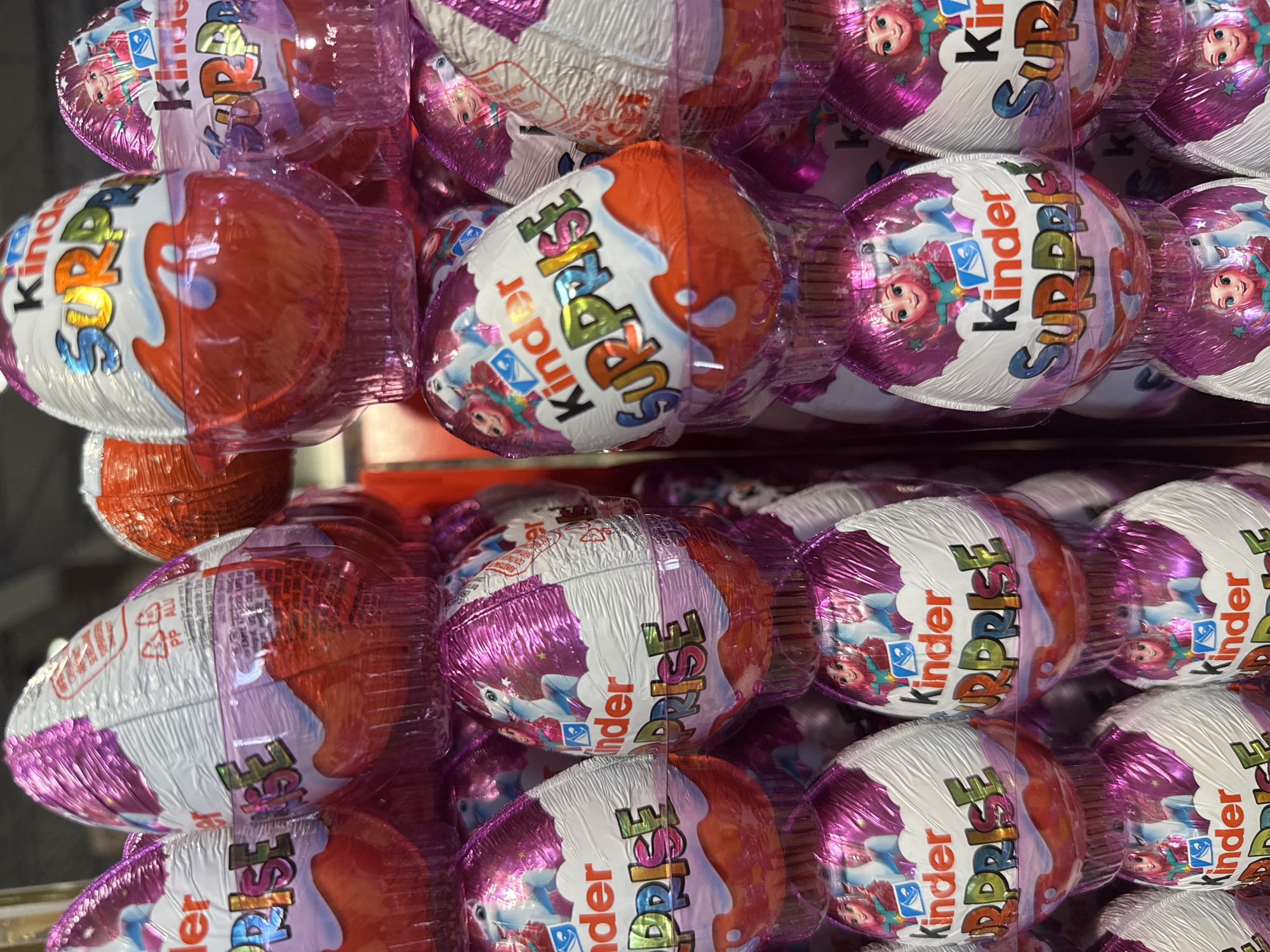 The image shows a close-up of a stack of Kinder Surprise eggs. The eggs are individually wrapped in foil, with half of each egg being white and the other half being either red or purple. The brand name "Kinder Surprise" is prominently displayed on the packaging. The environment suggests the image was taken at the city of Darien, in the United States of America.