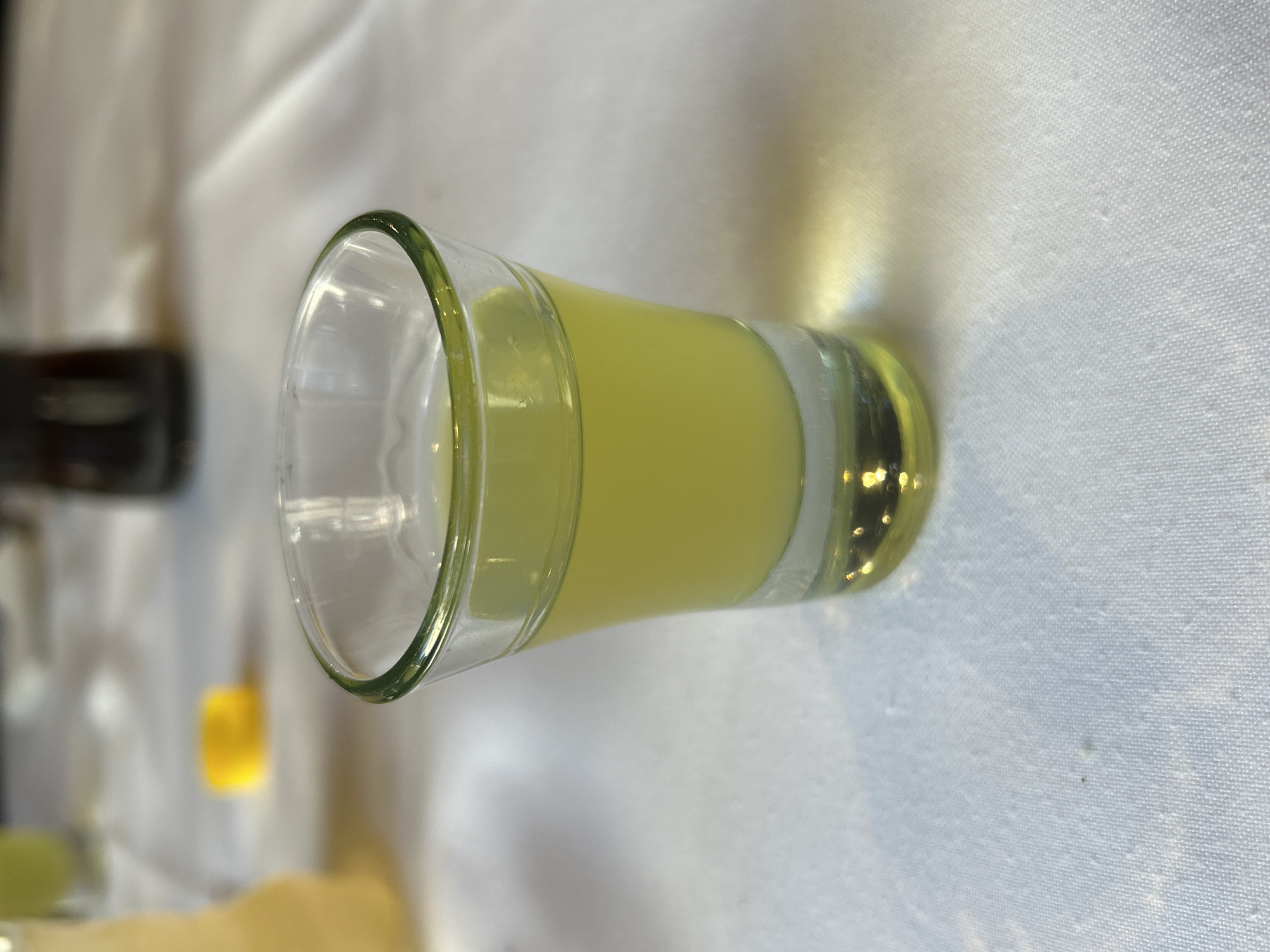 A close-up, slightly tilted, photograph shows a glass shot glass filled with a pale yellow liquid. The glass has a thick, green rim. It rests on a textured white surface. In the blurred background, other indistinct objects are present, including what appears to be a dark bottle, and a yellow object. The location and city of Buenos Aires, Argentina cannot be determined from the image.
