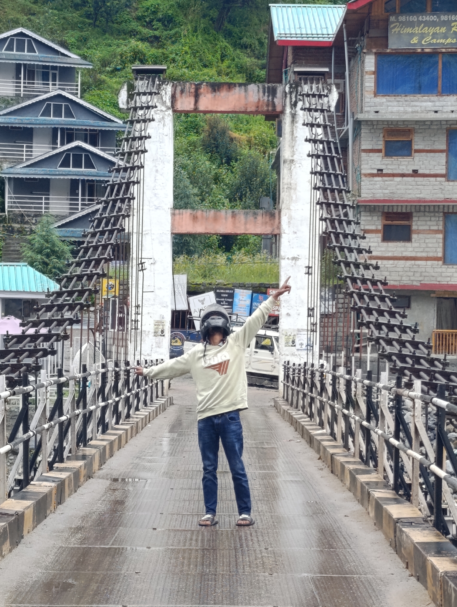 A full shot captures a person on a bridge in Kalath, India. The individual, wearing a light-colored hoodie and blue jeans, stands with arms outstretched, pointing upwards with their left hand. They are also wearing a helmet and sandals. The bridge is a metal suspension bridge with a wet, patterned surface. On either side of the bridge, there are railings and a stone wall. In the background, to the left, are multiple stacked buildings with blue roofs and white balconies. To the right, a building with brick and stone facade is visible, with signage that reads "M. 98160 43400, 98160... Himalayan Resorts & Camps." The environment appears to be a mountainous region with green foliage visible on the slopes behind the buildings. The sky is overcast, and there are visible water puddles on the bridge surface, suggesting recent rain.
