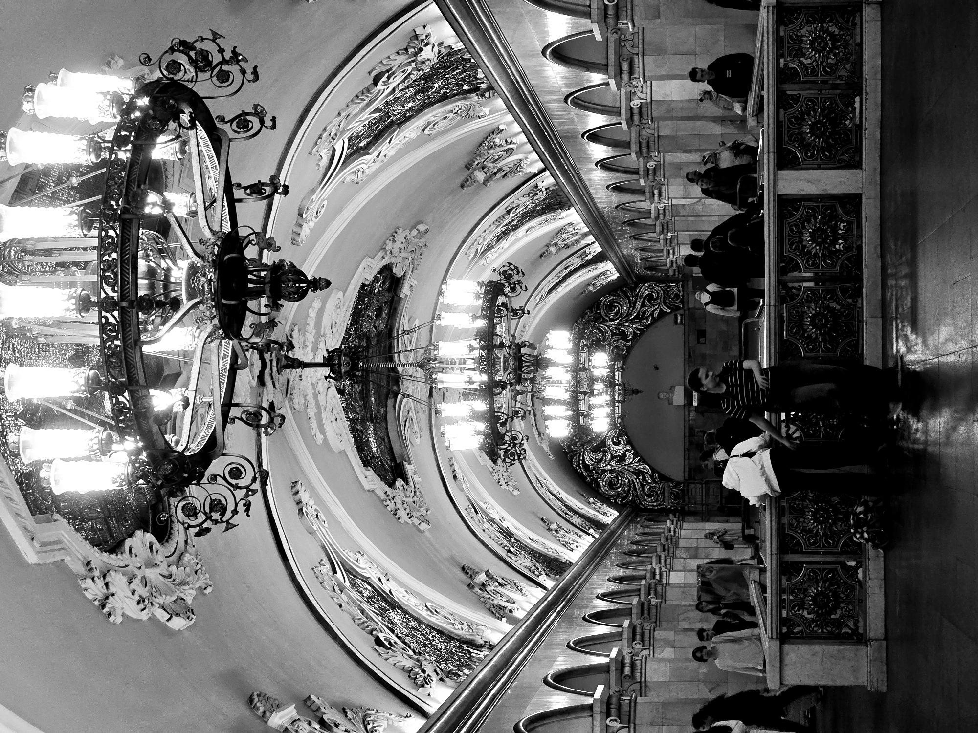 A black and white, upside-down, eye-level shot depicts a hallway in a location in the city of Unknown, Unknown. The ceiling features a curved design with intricate artwork and ornate lighting fixtures. Chandeliers hang at regular intervals. On the right side of the frame, people stand and walk along a walkway. The walkway is adorned with decorative metalwork and marble. The photograph is taken from a low angle, emphasizing the height and grandeur of the hallway.