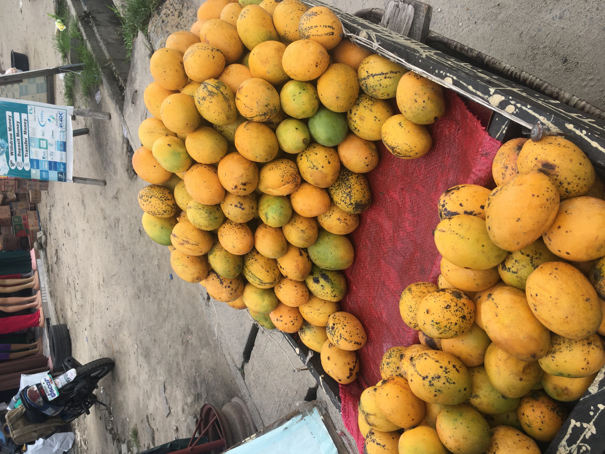 The image shows a large pile of mangoes on a cart, with some ripe and yellow, and others still green. The cart is lined with red fabric and has a weathered wooden frame.
To the left, there is a sign advertising money transfer services, displaying logos of various financial institutions and brands. A motorcycle is partially visible on the ground to the left of the cart, and there is a glimpse of clothing and beds in the bottom left corner.
The scene appears to be a street in Ado, Nigeria, with the mangoes being sold from a cart. The presence of the money transfer sign suggests a bustling commercial area.
