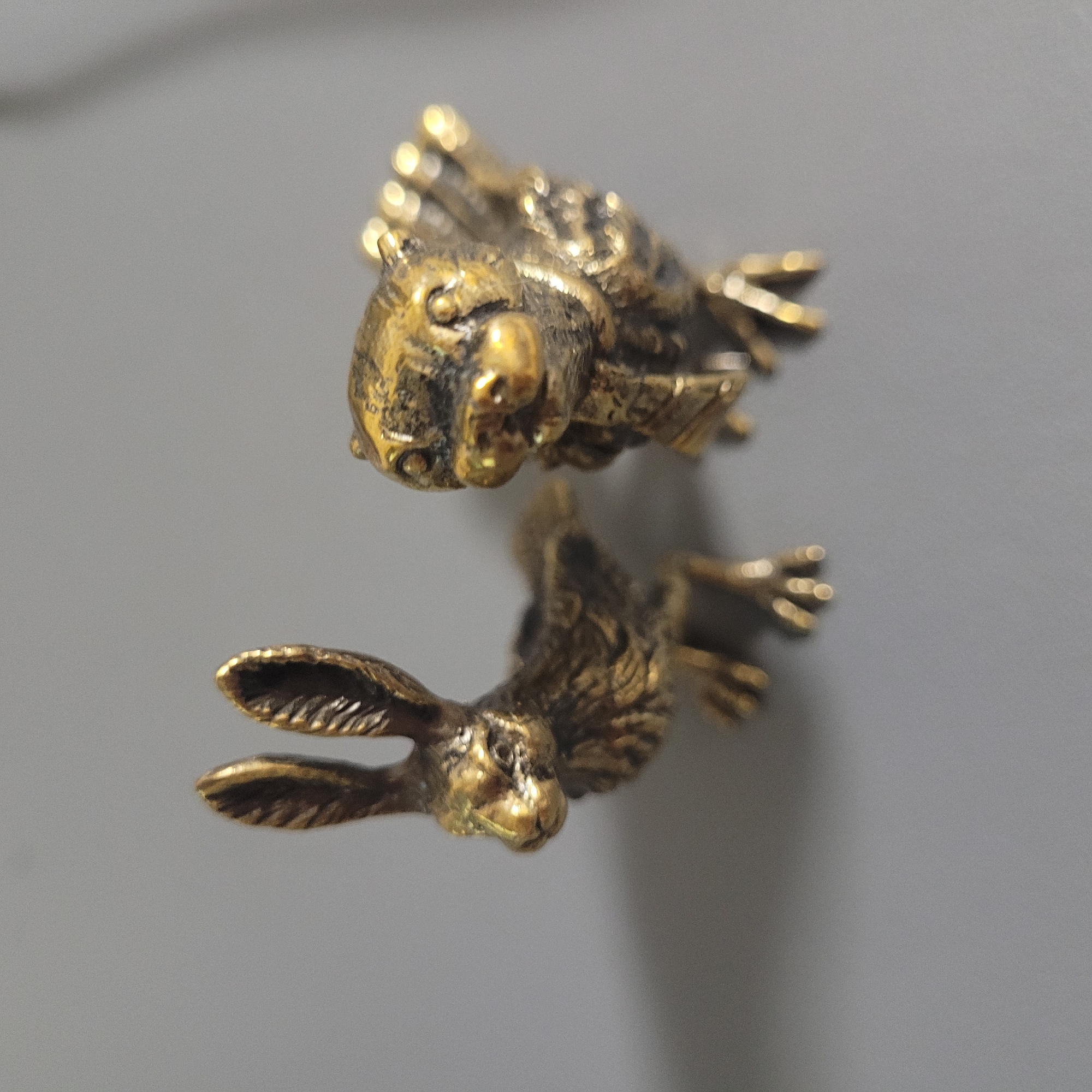 Two small, ornate bronze figurines of animals are positioned on a plain, grey surface. The figurine on the left depicts a rabbit, facing left in profile. It has long, upright ears with visible texture lines and detailed features on its face, including its nose and eyes. Its body is rendered with fur texture, and it stands on four legs. The figurine on the right is a stylized depiction of a bear or similar creature, facing forward and slightly to the right. It has large, round eyes, a prominent snout, and detailed paws. Its body also exhibits a textured surface, and it appears to be wearing a decorative collar or harness. The lighting casts subtle shadows, highlighting the metallic sheen of the bronze. The background is a uniform, soft grey, devoid of any discernible features. The scene is static and does not depict any action or interaction.