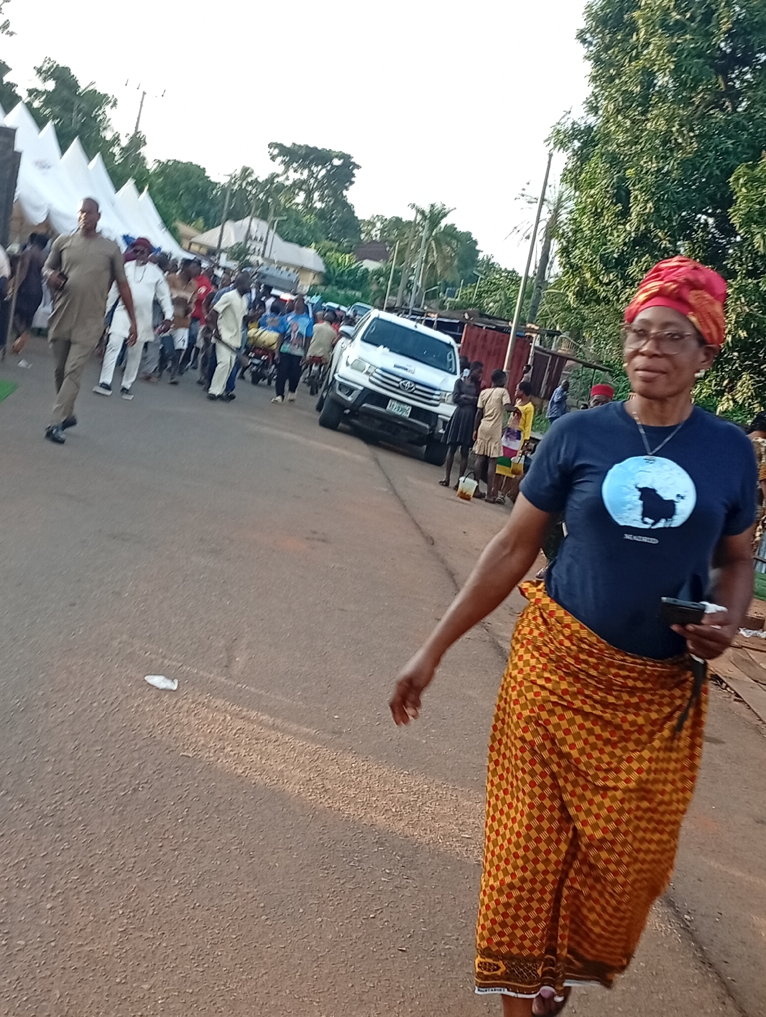 A medium shot captures a woman walking forward on a paved road in Nsugpe, Nigeria. She is wearing a dark blue t-shirt with a circular graphic of a bull, a red and yellow patterned wrapper, and a red headscarf. She is also wearing glasses and holding a mobile phone in her right hand. In the background, several people are visible, along with white tents, a white SUV, and trees. The road is lined with trees and some buildings. The overall scene appears to be an outdoor event or gathering.