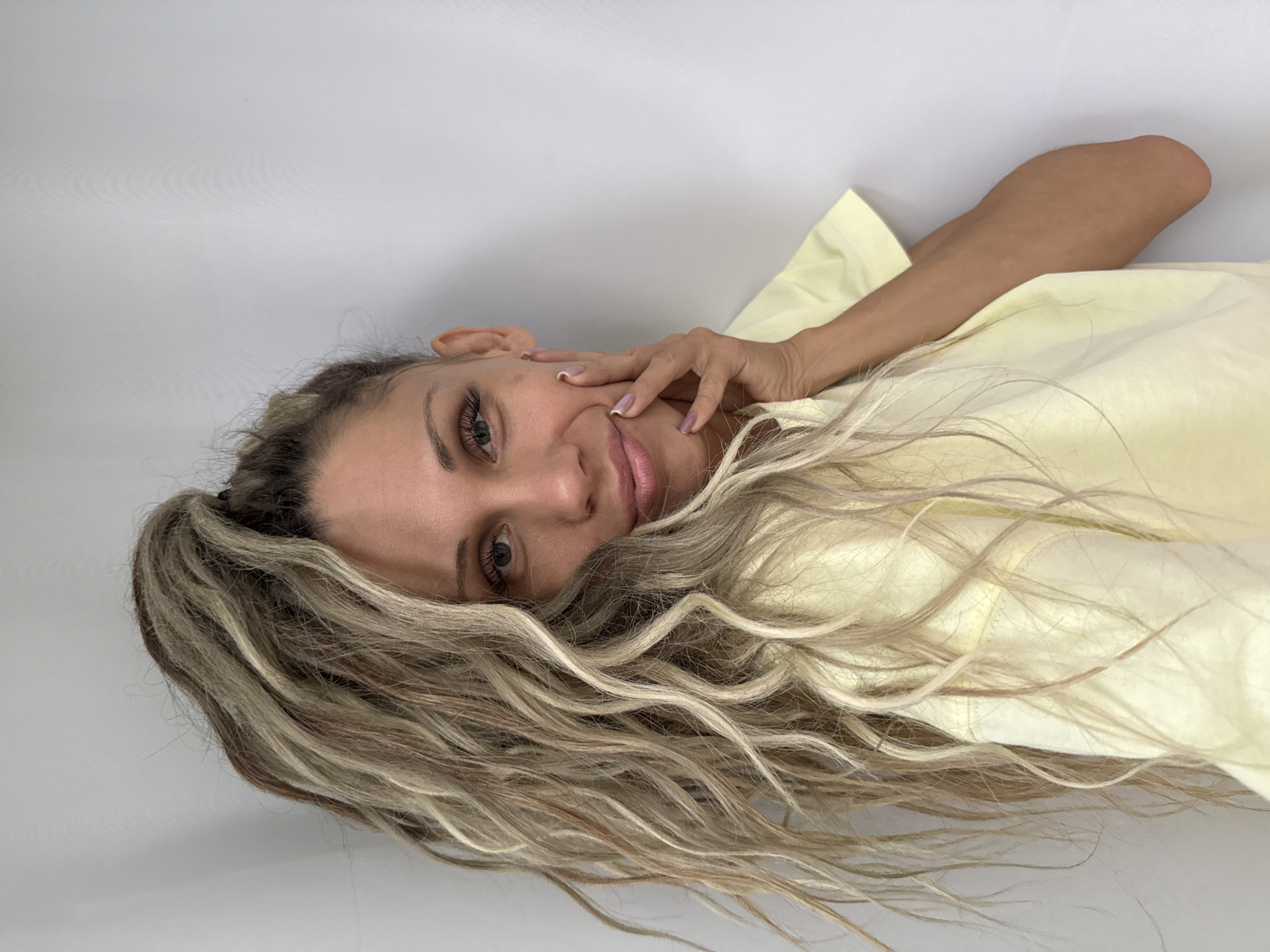 A woman with long, blonde, curly hair pulled up in a high ponytail is seen from the chest up, with her left hand touching her face. She is wearing a light yellow t-shirt and is against a plain, light-colored background. The lighting is soft, casting a gentle glow on her face. Her eyes are looking towards the camera, and she has a slight smile.