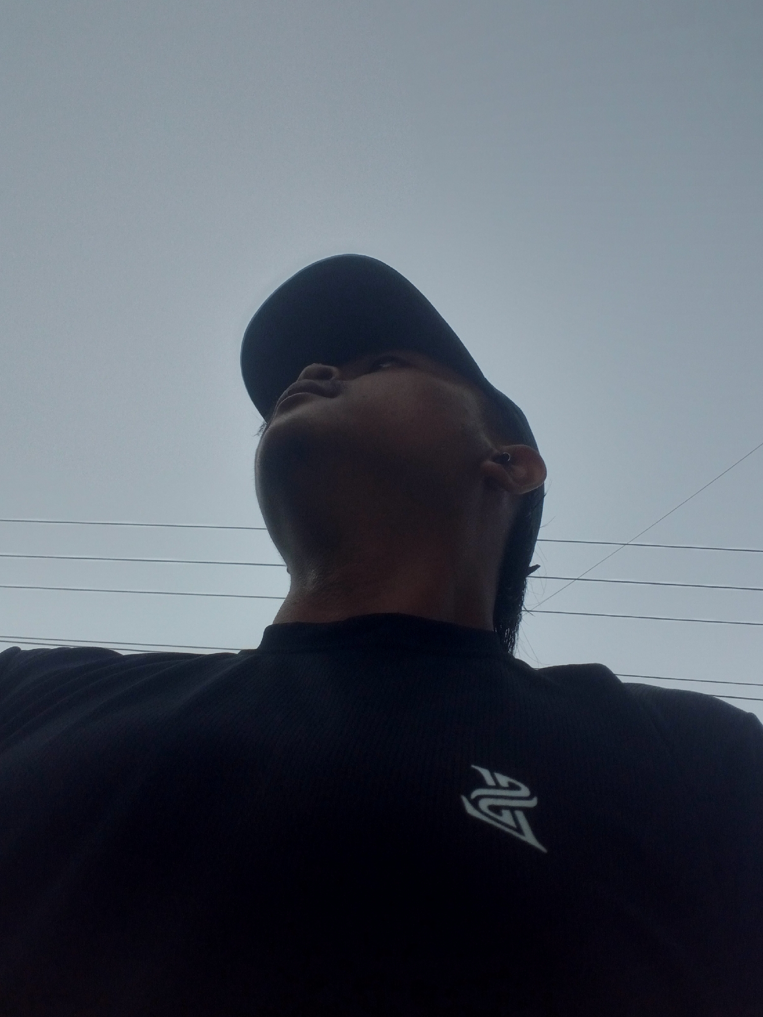 The image, taken from a low angle, shows the upper body of a person wearing a black t-shirt with a white logo. The person is also wearing a dark baseball cap, and is looking upwards. Their ear is visible and appears to have an earring. In the background, there are several horizontal power lines against a grey, overcast sky. The image does not contain any specific visual cues that would indicate the location is Kubang Jaya, Indonesia.
