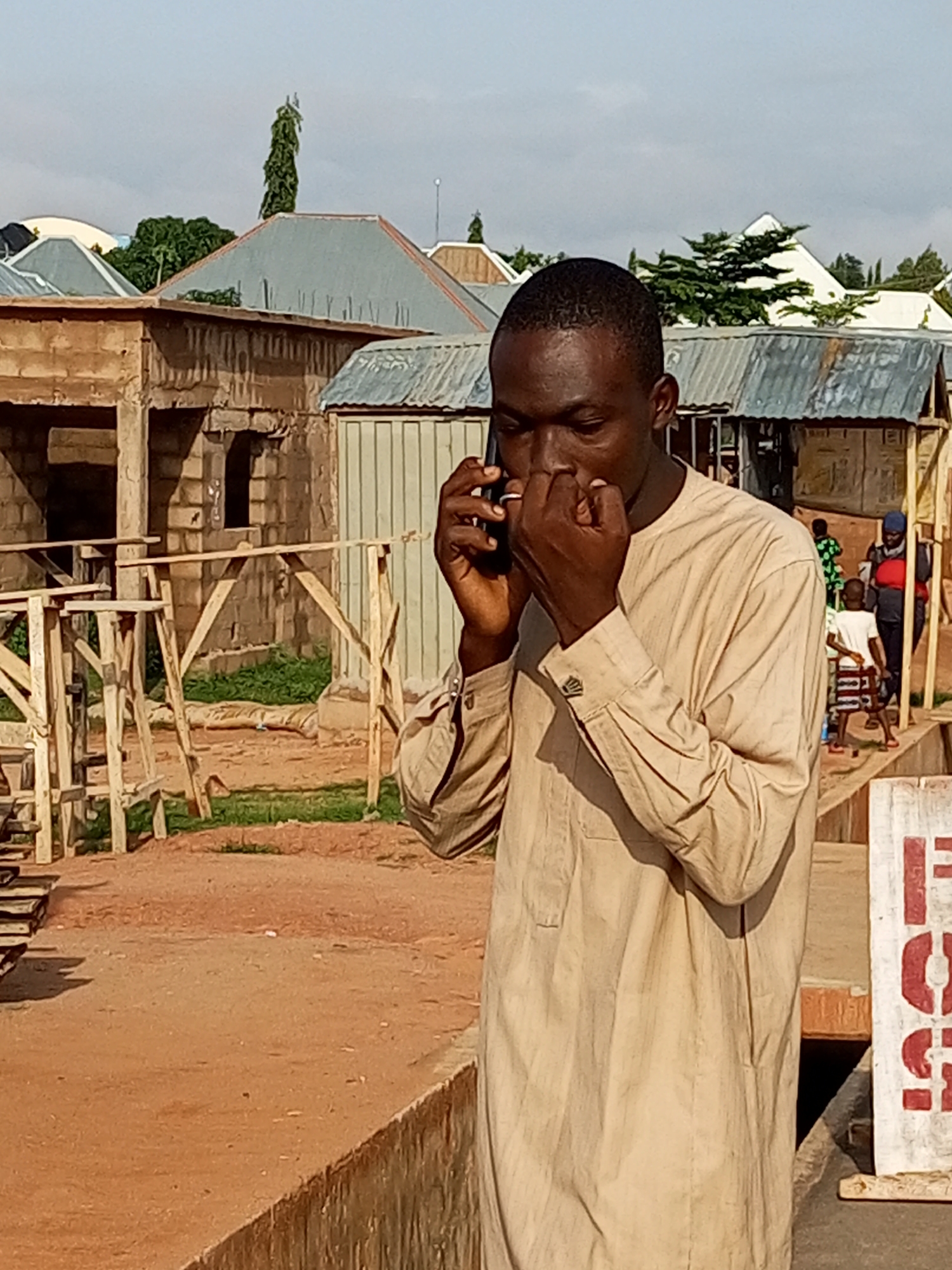 A man is standing outside, holding his phone to his ear and talking. He is wearing a light brown, long-sleeved shirt and light brown pants. He is looking down at his phone. In the background, there are several buildings with corrugated metal roofs. There is also some construction scaffolding made of wood. The ground is covered in dirt and there is some green grass. The sky is blue with some clouds. The location is in the city of Tempure, Nigeria.