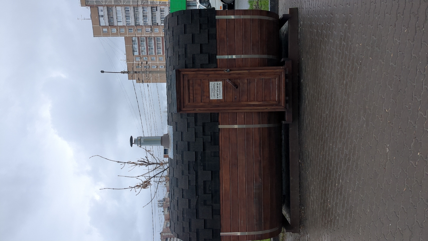 This media file captures a barrel-shaped wooden sauna or bathhouse, commonly known as a "banya," positioned outdoors on a paved surface. The structure is constructed from dark wooden planks with visible metal bands, and its curved roof is covered with black shingle tiles. A prominent silver-colored metal chimney extends upwards from the top.
A small, rectangular wooden door is visible on the side of the barrel structure. Attached to this door is a white sign with black Cyrillic text. The text clearly states "БАНЯ" (BANYA, meaning bathhouse or sauna) and a phone number: "8-951-272-89-37". Below the phone number, there appears to be additional text, possibly "Бочки" (Boats/Barrels), likely referencing the barrel design.
The setting is an urban or suburban environment. The ground consists of damp, interlocking grey-brown pavers. In the background, several multi-story residential buildings with numerous windows can be seen under an overcast sky. Utility poles and a network of overhead power lines are present, stretching across the upper part of the image. Bare tree branches are visible near the chimney. The cloudy sky and damp ground suggest a cool, possibly recently rainy, daytime. There are no people visible, and no specific activity is occurring. The visible Cyrillic text and architectural style indicate a location in Russia, specifically Ulyanovsk as per the provided context.