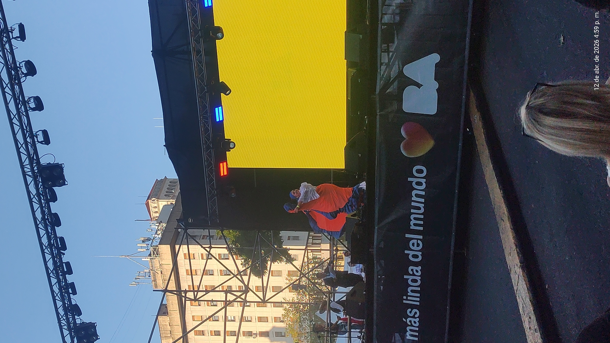 The image shows a stage set up in what appears to be Buenos Aires, Argentina. In the foreground, part of the event stage is visible. It features a yellow LED screen and the slogan "BA más linda del mundo" (BA most beautiful in the world).
Two dancers are captured mid-performance on stage, wearing traditional costumes.
In the background, there is a building and blue sky. The image also shows stage lighting equipment.
In the upper-right corner, there's a time stamp indicating "12 de abr. de 2026 4:59 p. m." (April 12, 2026, 4:59 p.m.).