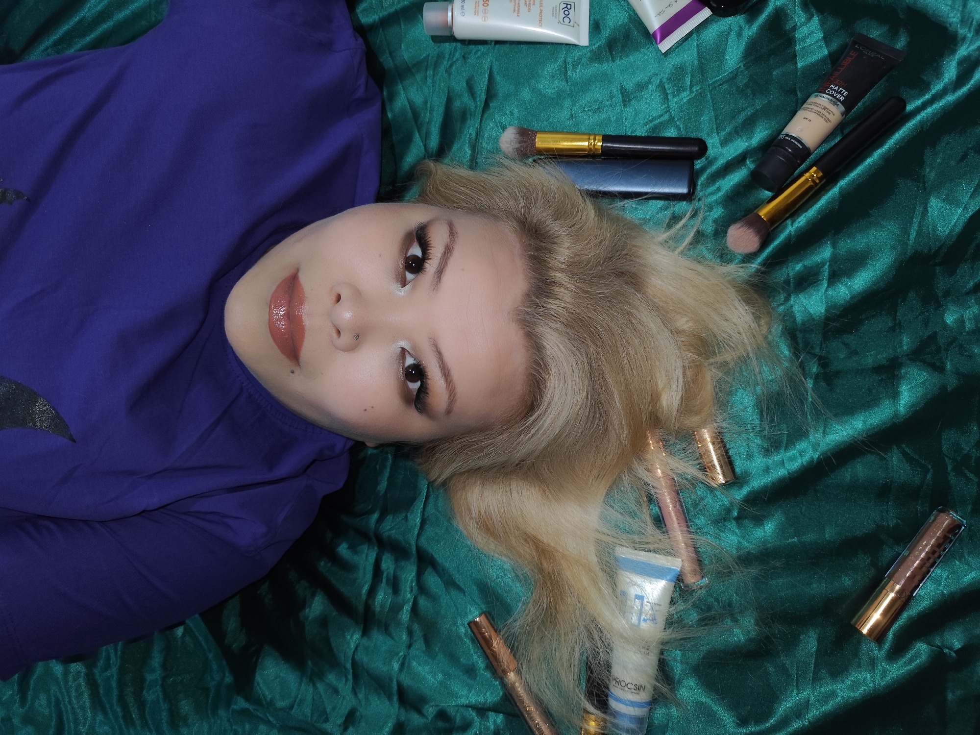 The image shows a young woman lying on a dark green, wrinkled fabric, surrounded by various cosmetics and beauty products. She is wearing a purple t-shirt and has blonde hair. Her makeup includes light eyeshadow, lipstick, and mascara. Several cosmetic tubes and brushes are arranged around her head on the fabric.
While the city of Sarıyer, Turkey, is mentioned, there's no information in the image that confirms this location.