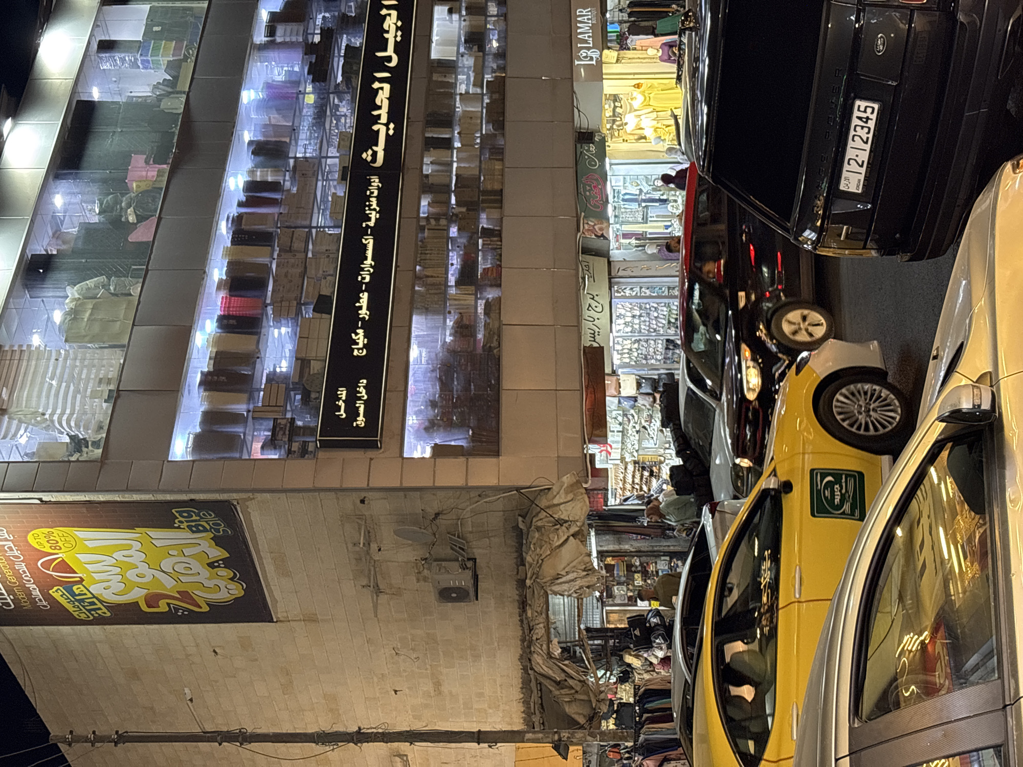 The image captures a street scene in Amman, Jordan, at night. In the foreground, a black Range Rover with a Jordanian license plate reading "12-12345" is parked on the right. To its left, a yellow taxi is visible, with a green emblem on its side. Several other cars, including a black SUV with a red roofline, are also present in the street, suggesting traffic congestion.
The background features a multi-story building with large glass windows showcasing various products on shelves. A prominent banner on the left side of the building advertises a sale with "UP TO 80% OFF". The building's facade is illuminated, highlighting the display windows and signage. Below the main building, there are smaller shops and stalls with clothing and other merchandise on display, with a few people browsing. The street is paved, and a utility pole is visible on the left side of the image. The overall atmosphere is that of a busy urban commercial area at dusk or night.