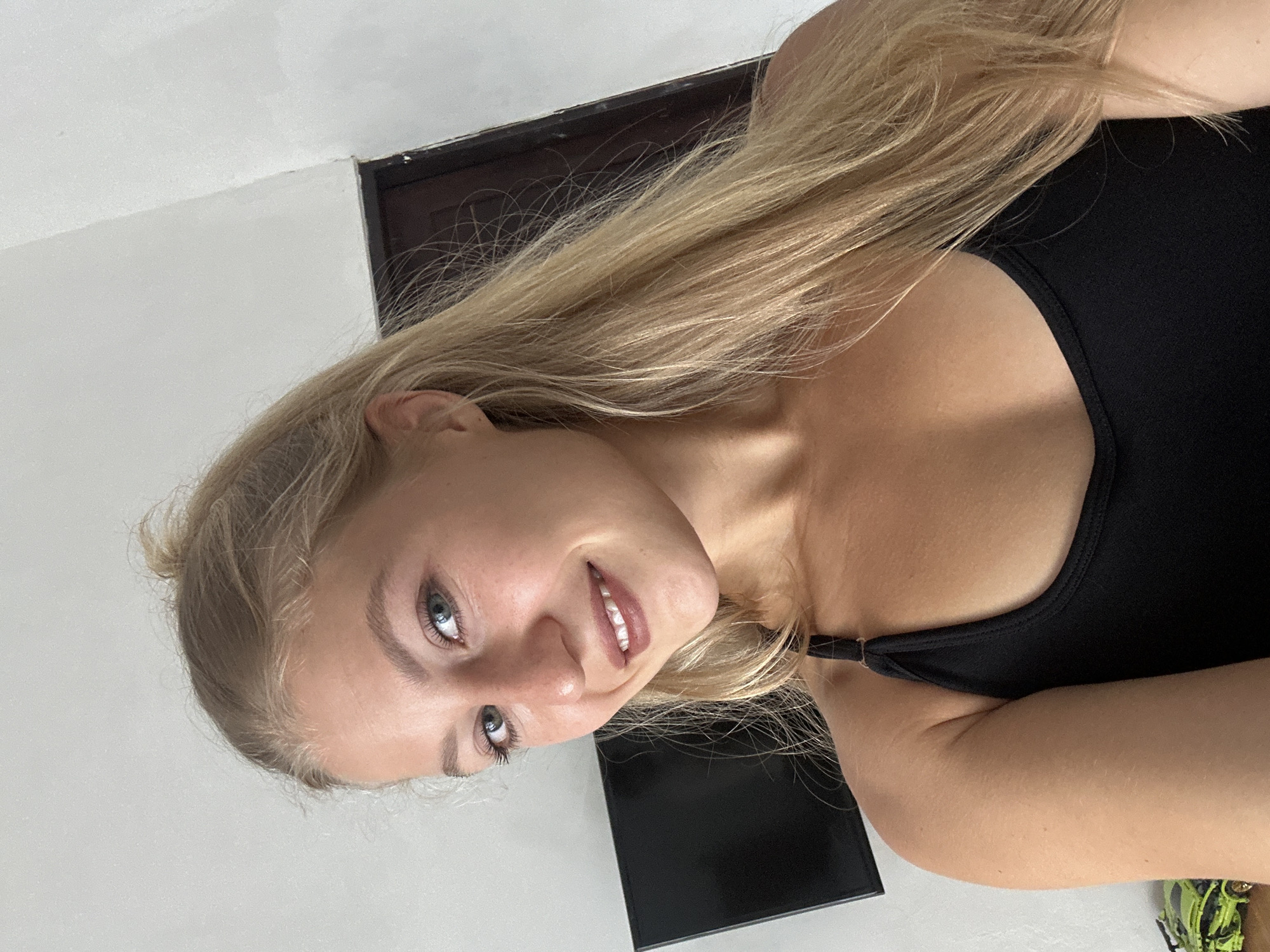 A headshot-style portrait captures a young woman looking directly at the camera with a soft, engaging smile. She has long, straight blonde hair that flows over her shoulders and a healthy, tanned complexion. Her prominent blue eyes are highlighted with what appears to be light makeup, including mascara, and her lips wear a subtle color. She is dressed in a simple black tank top or camisole.
The background is minimalistic, primarily consisting of a plain white wall, indicative of an indoor setting. In the upper right portion of the image, a dark wooden frame is visible where the wall meets the ceiling, suggesting a door or window frame. A dark, rectangular object, possibly a television or piece of furniture, is partially visible in the background to her lower right. A small detail of a green and black patterned item can also be seen in the bottom right corner, perhaps part of her clothing or an object nearby.
The lighting is bright and even, illuminating her face and hair. She conveys a pleasant and relaxed emotion. There is no visible text in the image. The location context for this media file is Kuta Selatan, Indonesia.
