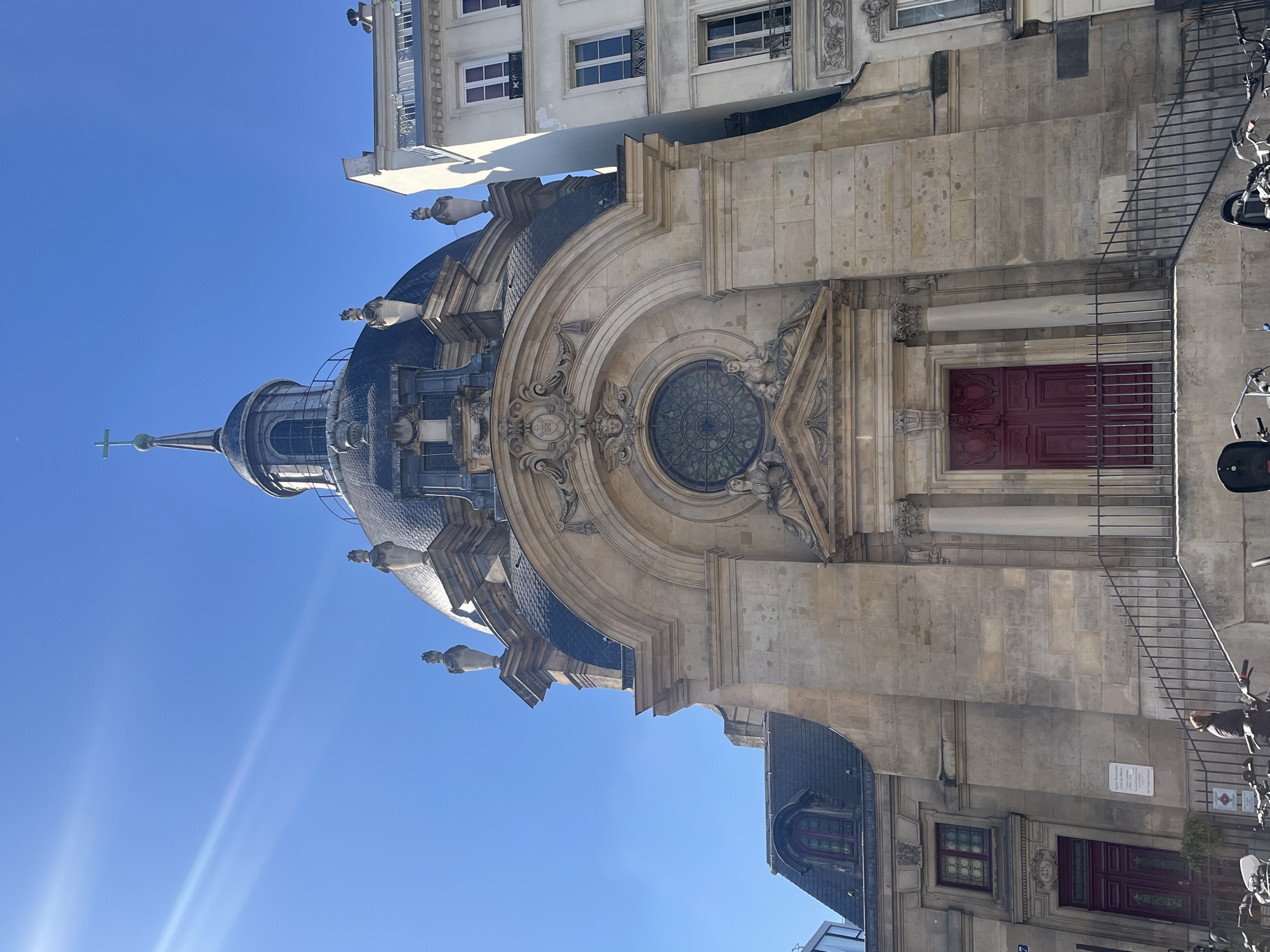The image presents a striking view of an ornate, historic building, likely a church or chapel, under a brilliant clear blue sky. The main subject is characterized by its prominent dark-tiled dome, adorned with several grotesque gargoyle-like statues. A slender spire rises from the dome, topped with a cross, reaching towards the sky. The building's facade is constructed from light-colored stone, featuring elaborate architectural details including columns, cornices, and intricate carvings that frame a large, circular stained-glass window. Below this decorative window, a set of deep red double doors serves as an entrance.
To the right of the main structure, a black metal railing runs along the sidewalk, where several motorcycles or scooters are parked. A person, partially visible, stands near these parked vehicles. Parts of other buildings are also visible: an apartment-style building with white-framed windows is seen in the upper background, and a lower building with a dark roof and a smaller, arched window is present in the foreground. The bright, sunny weather is evident from the strong sunlight and a noticeable lens flare in the upper left of the sky.
A white plaque attached to the wall near the parked scooters displays French text. It reads: "Ce fut la / Maison de la famille / DE MEDICIS / Avant d'habiter le / Palais des Tournelles." This translates to: "This was the / House of the DE MEDICIS family / Before living in the / Palais des Tournelles," indicating a significant historical landmark in Paris, France. The overall scene captures a sunny day in an urban setting, showcasing impressive European architecture.