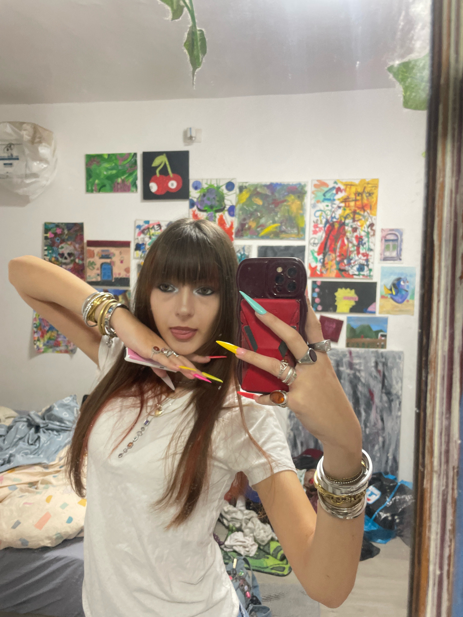 This is a medium shot photograph taken in a bedroom. A young woman with long brown hair and bangs is standing in front of a mirror, taking a selfie. She is wearing a white t-shirt and has long, brightly colored acrylic nails. She is adorned with several bracelets on her left arm and rings on her fingers. The bedroom walls are covered with various paintings and artwork. A laundry bag is visible in the background, as well as a bed with rumpled sheets. The image is not identifiable as being located in the city of Raanana, Israel.