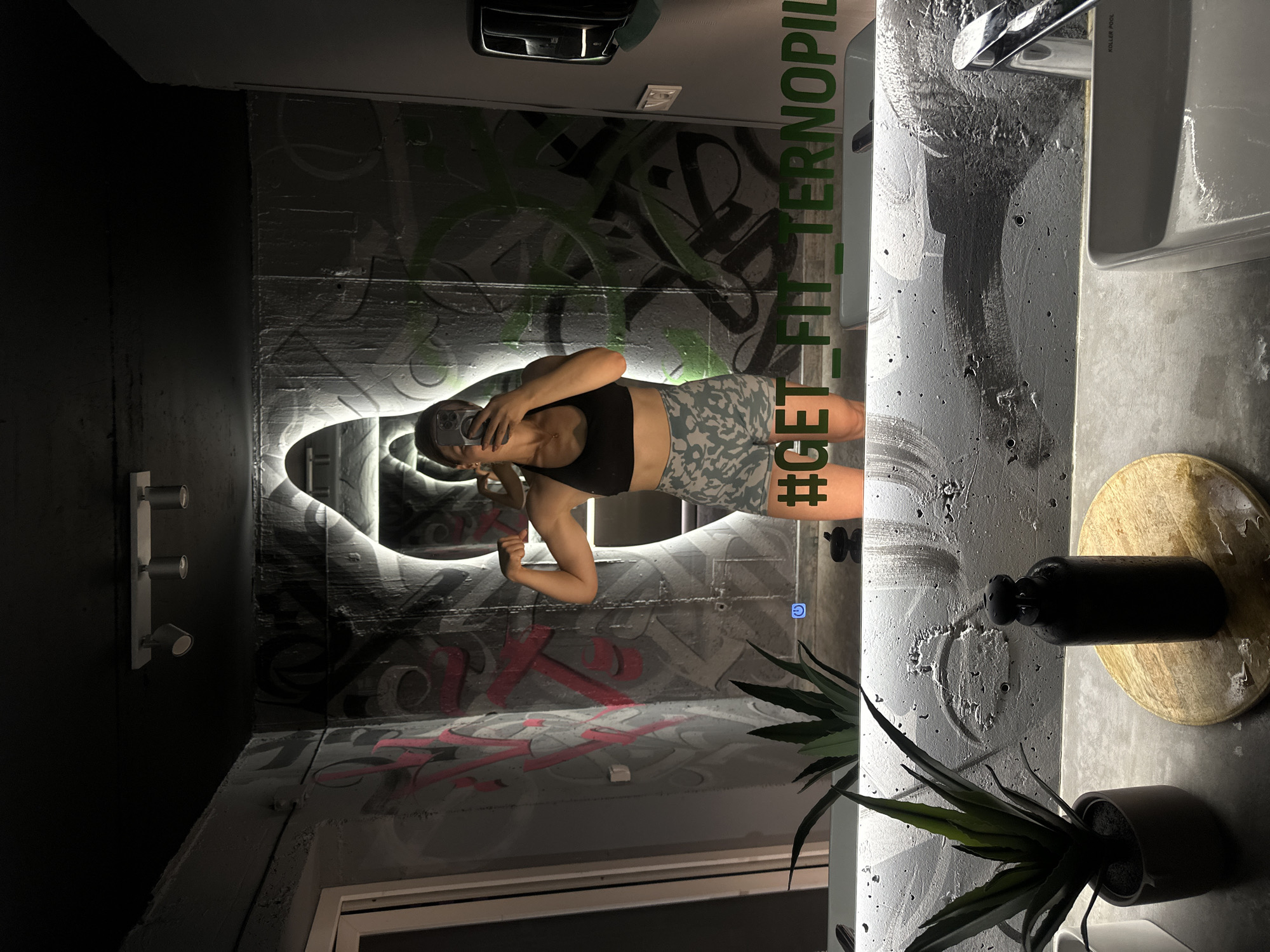 A woman is taking a mirror selfie in a modern restroom with graffiti art on the walls. She is wearing a black sports bra and camouflage shorts, flexing her bicep. The mirror has the hashtag "#GET_FIT_TERNOPIL" etched into it. The restroom features a concrete countertop with a sink, a black water bottle, and a potted plant. The lighting is dim, with spotlights on the ceiling and a backlit mirror. The graffiti art on the walls appears to be in shades of gray, black, and pink.