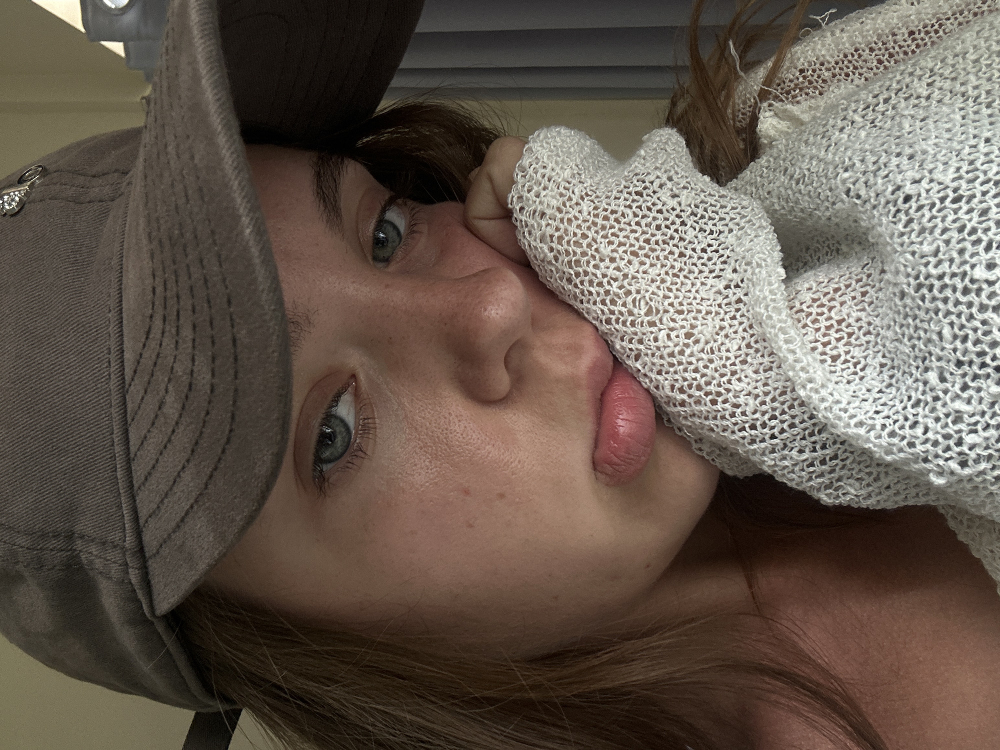A close-up shot focuses on the left side of a young woman's face, wearing a brown baseball cap and a white, loosely knit top. She rests her left hand, covered by the top's fabric, against her cheek. Her blue eyes are looking slightly to the left, and her lips are slightly parted. The background shows a plain wall and a curtain. The scene appears to be indoors, possibly in a home, but no specific location in a city can be determined.