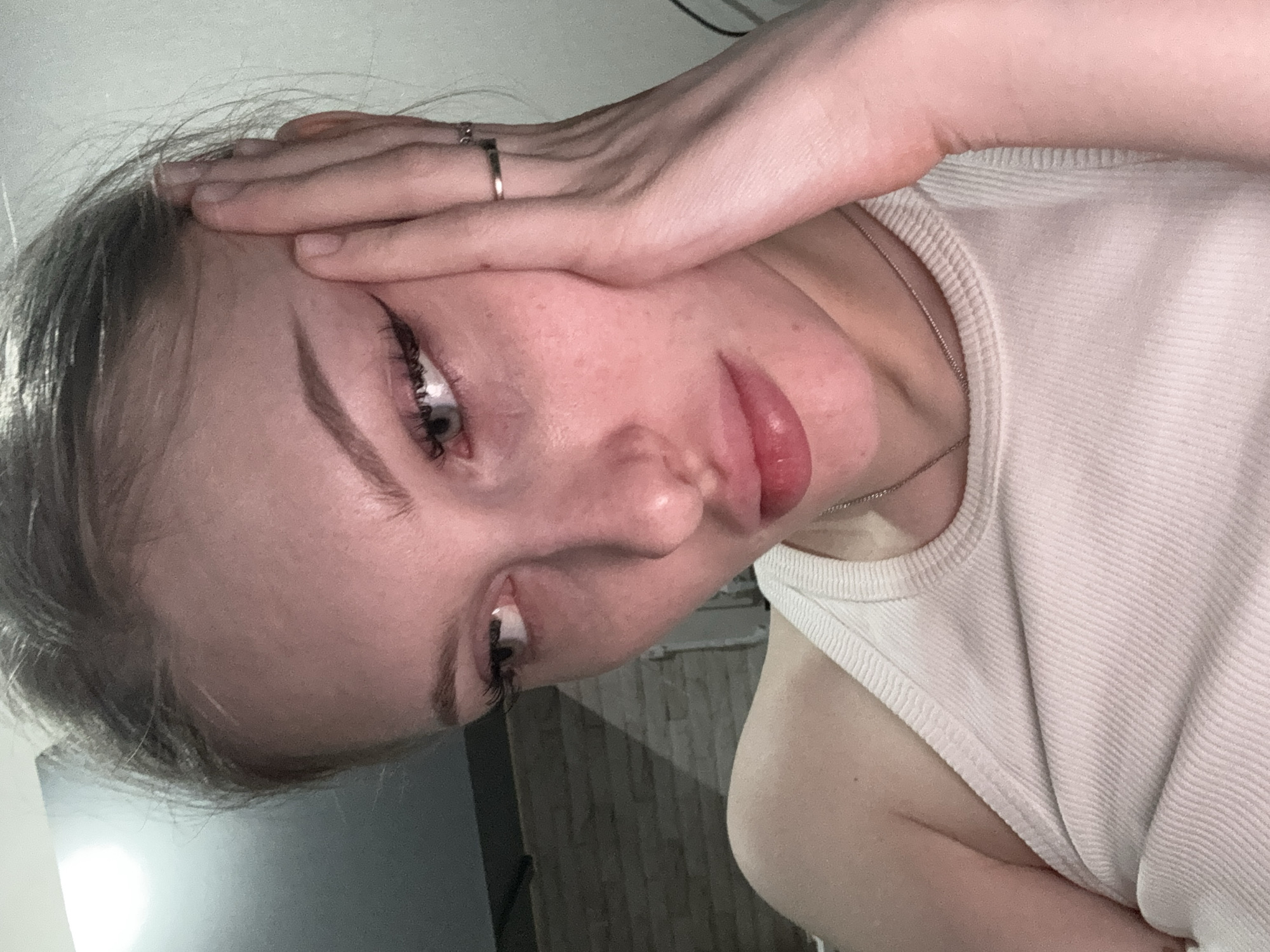 A close-up, slightly out-of-focus, selfie portrait shows a young, light-skinned person with light-colored hair. The person is wearing a white tank top and a ring is visible on the hand that is positioned on the forehead. The background is blurred, showing a dark wall and what appears to be an edge of a light-colored surface. The picture might have been taken indoors. There is no information to confirm this was taken in Perm, Russia.