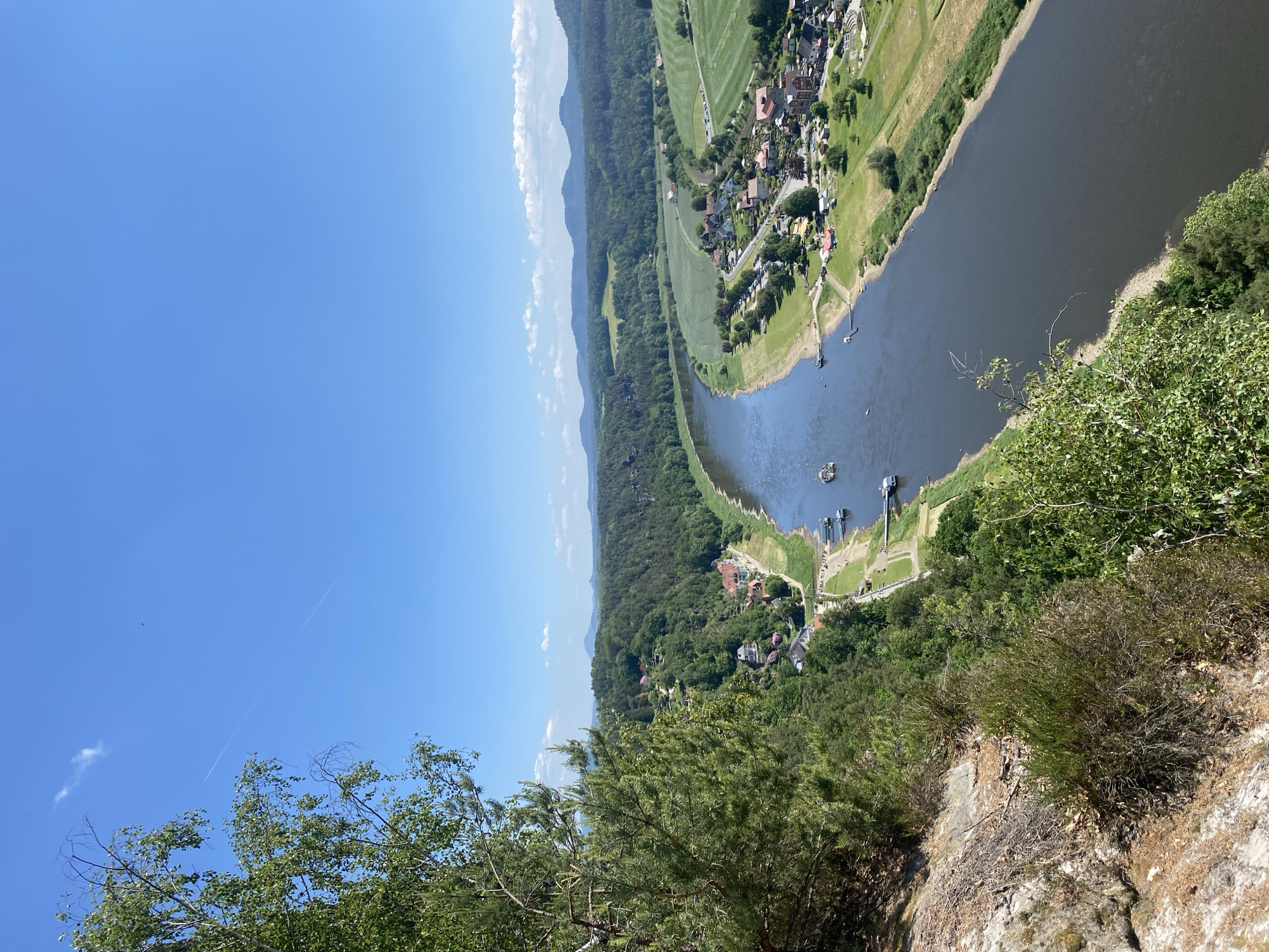 The image shows a view of the Elbe River from the Bastei rock formation in Lohmen, Germany. The river flows in a wide bend, with a town visible on the opposite bank. The riverbank features vegetation, and several small boats are moored along the shore. The sky is blue with a few scattered clouds. The foreground includes foliage and rock formations, giving the impression of looking out from a high vantage point.