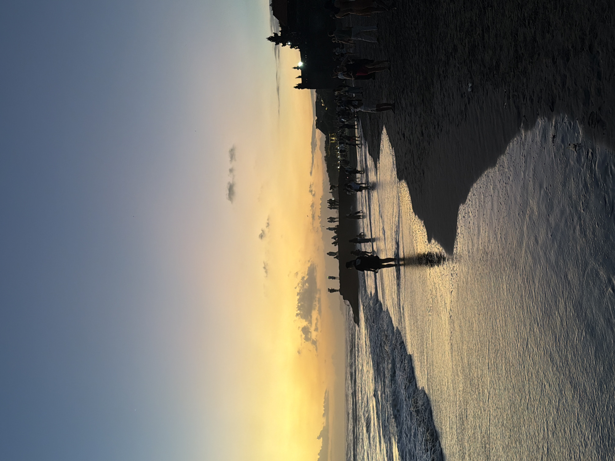 This is a photograph of a beach in North Kuta, Indonesia, during sunset. The sky is a gradient of deep blue at the top transitioning to a warm yellow and orange near the horizon, with a few scattered clouds. The sun is setting over the ocean, casting a golden glow on the water and the wet sand. Gentle waves are rolling onto the shore.
There are numerous people on the beach, most of them appearing as silhouettes against the bright sky. Some are walking along the shoreline, while others are standing on a rocky outcrop or near buildings in the background. The buildings, possibly temples or resorts, have distinctive architectural features that suggest a Southeast Asian setting. The sand is dark and appears to be a mix of fine grains and possibly volcanic ash, which is common in some Indonesian beaches. The overall atmosphere is one of peaceful recreation and natural beauty as the day concludes.