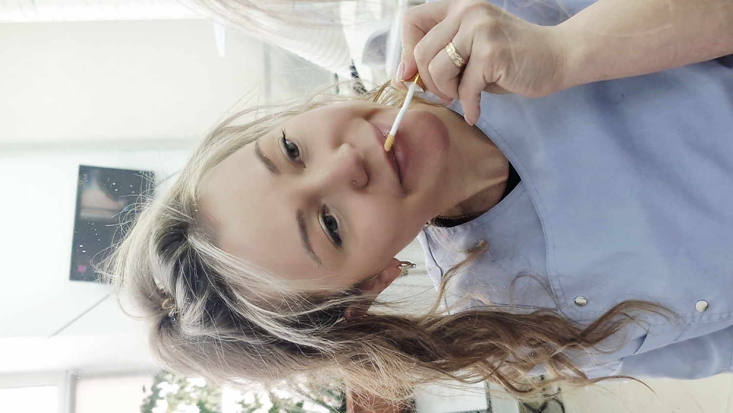 The image, rotated counter-clockwise, depicts a woman applying a product to her lips. She has long, wavy blonde and light brown hair, which frames her face. She is wearing a light blue, possibly uniform-style smock or top, featuring metallic snap buttons on the shoulder.
The woman is shown from the chest up, with her head slightly tilted. Her right hand, adorned with a gold-toned ring on her ring finger, holds a white applicator with a golden cap. The applicator, which appears to have a yellowish, viscous product on its brush tip, is touching her upper lip. Her lips appear full. She has a soft, slight smile and looks directly towards the viewer with open eyes.
The background is bright, suggesting an indoor setting like a beauty salon, clinic, or studio. Reflections in what appears to be a large mirror show a clean, light-colored wall and a dark, rectangular object, possibly a screen or framed picture, further in the background. The scene suggests an activity related to beauty care, likely the application of lip gloss, balm, or post-treatment care. There is no visible text in the image.