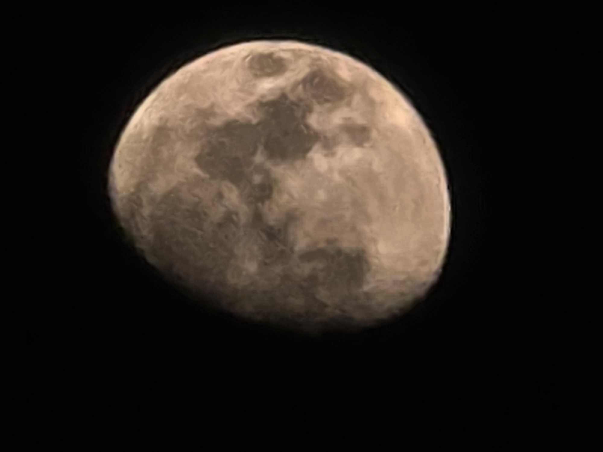 The image is a close-up, somewhat blurry photograph of the moon against a black background. The moon is illuminated, showing surface features such as craters and maria, and appears to be in a gibbous phase. The color of the moon is a warm, light brown or tan. There is no discernible context related to the city of Yambol, Bulgaria, or any urban environment present in the image.
