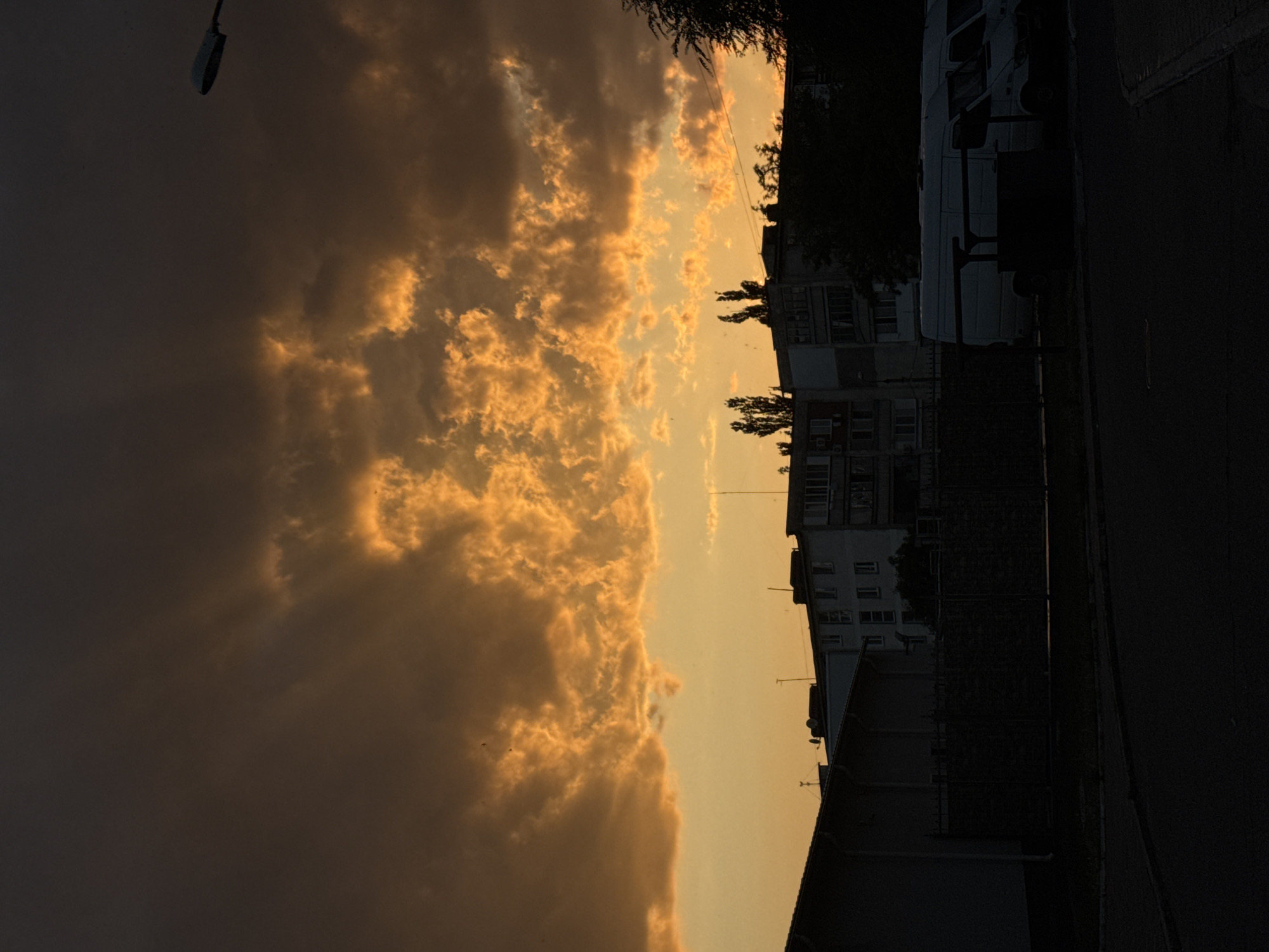 The image depicts a twilight scene in Tiraspol, Moldova, with a sky dominated by dramatic, brightly lit clouds in hues of orange and yellow against a darker, approaching night sky. In the foreground, a dark asphalt road curves towards the right, with a silhouetted fence running along the middle ground. Behind the fence, several white vans are parked, their forms indistinct in the low light. Further back, silhouetted apartment buildings rise, their windows reflecting the fading light. Trees, also silhouetted, are scattered amongst the buildings and on the right side of the frame. A street lamp is visible in the upper right corner. There are no visible people in the image.