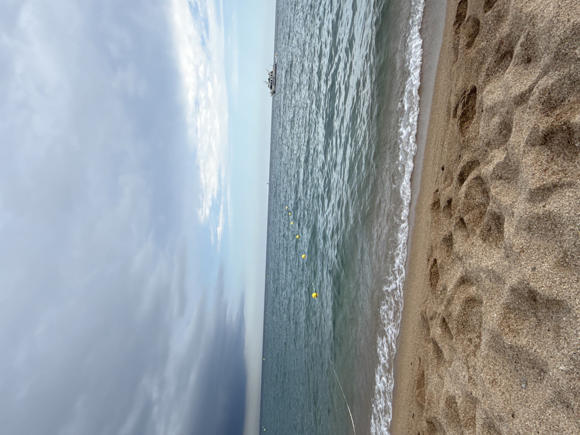 The image depicts a coastal scene on a sandy beach, likely in Blanes, Spain, given the context. The foreground is dominated by textured sand with footprints. The middle ground features the gentle lapping of waves on the shore and a rope with yellow buoys extending into the sea. In the distance, a white boat with "NEPTUNE" visible on its side is sailing on the rippling, blue-green water. The sky is overcast with dramatic, dark clouds on the left, transitioning to lighter, scattered clouds and patches of blue sky on the right. The overall impression is of a potentially changing weather condition at the seaside.