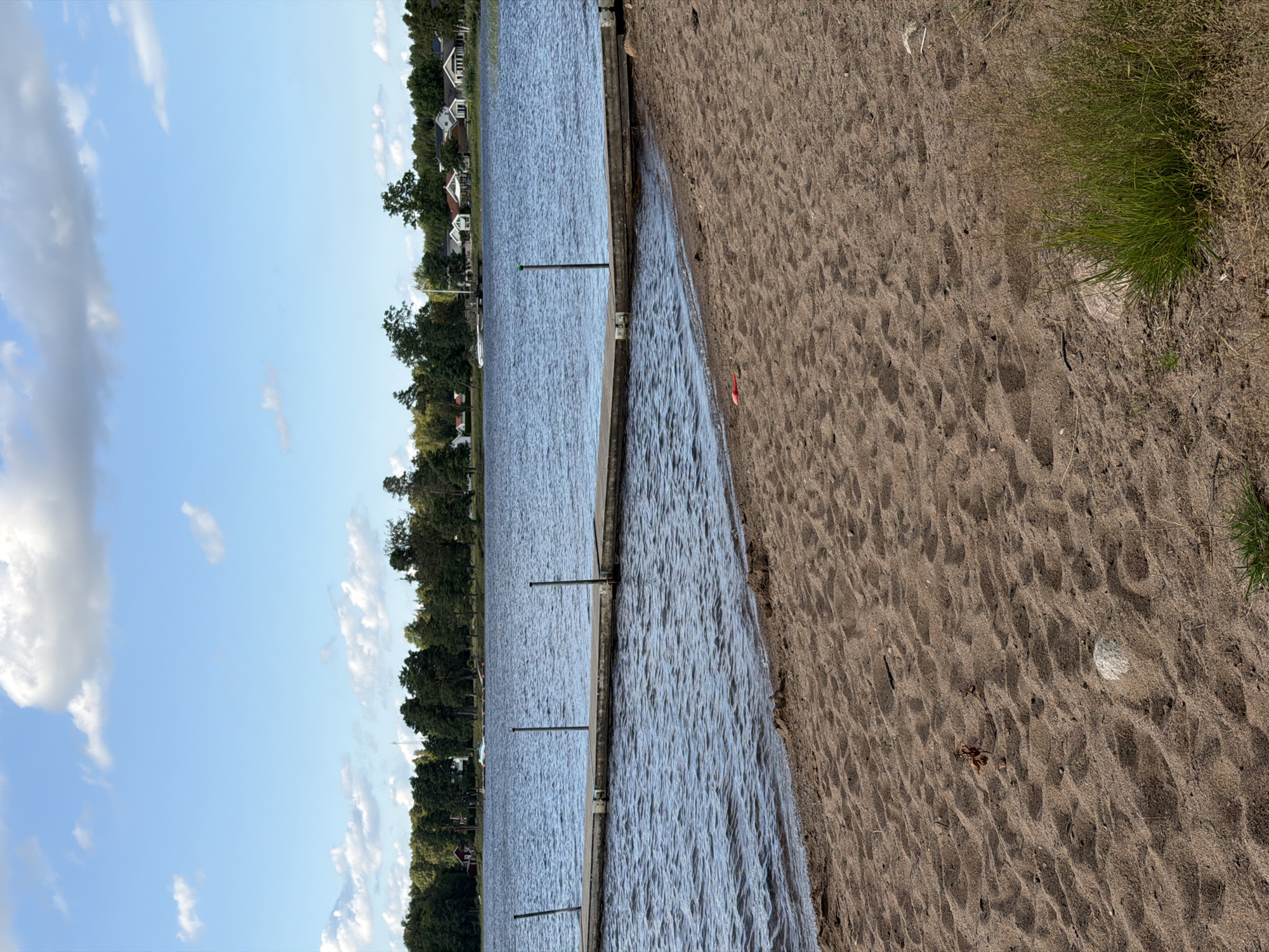 The image depicts a serene lakeside scene in Växjö, Sweden. The foreground is dominated by a sandy beach with scattered pebbles and a small cluster of green grass. The sand shows signs of footprints and some dried leaves. A red toy shovel is visible on the sand near the water's edge. A wooden jetty extends into the calm, rippling blue water of the lake. The middle ground features the lake and the jetty, with several vertical poles on the jetty. In the background, a line of trees borders the opposite shore, interspersed with several white and grey houses with red roofs. A few boats are docked in the water near the shore. The sky above is blue with scattered white clouds. The overall impression is one of a peaceful summer day at a lakeside location.
