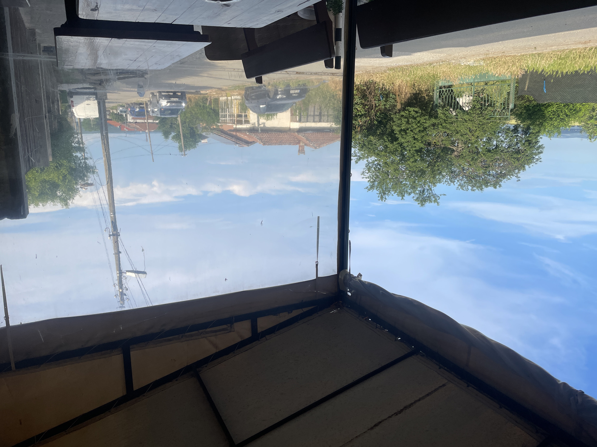 The image presents a detailed view from an outdoor covered seating area in Konstantinovo, Bulgaria, looking out onto a rural street and surrounding landscape on a clear day.
In the immediate foreground, dark wooden tables and chairs are visible, partially obscured. Above, a beige canopy structure supported by dark metal framing extends overhead, with clear plastic or vinyl screens serving as transparent walls, showing some smudges.
Outside, a dirt or gravel road runs horizontally across the midground. To the left, lush green vegetation, including trees, tall grasses, and weeds, lines the roadside. A decorative dark green metal gate, partly concealed by foliage, provides a break in the greenery. Beyond this, an expansive field or open area is visible, leading to distant hills under a bright blue sky with scattered white clouds.
In the center, a dark-colored SUV (appears to be a black Audi) is parked on a grassy verge, positioned in front of an older, single-story house featuring a red tile roof and light-colored walls. The house exhibits signs of age and is partially overgrown with vegetation.
To the right, a narrow, unpaved or semi-paved street extends into the distance. A tall wooden utility pole, equipped with multiple cross-arms and power lines, stands prominently by the roadside. Several vehicles are present on the street: a silver SUV is observed moving towards the viewer, while a blue truck and a white cargo van are parked further down the road. Additional houses with red roofs and numerous green trees line both sides of the street as it recedes into the background.
No people are visible within the frame, and the scene is illuminated by strong daylight.