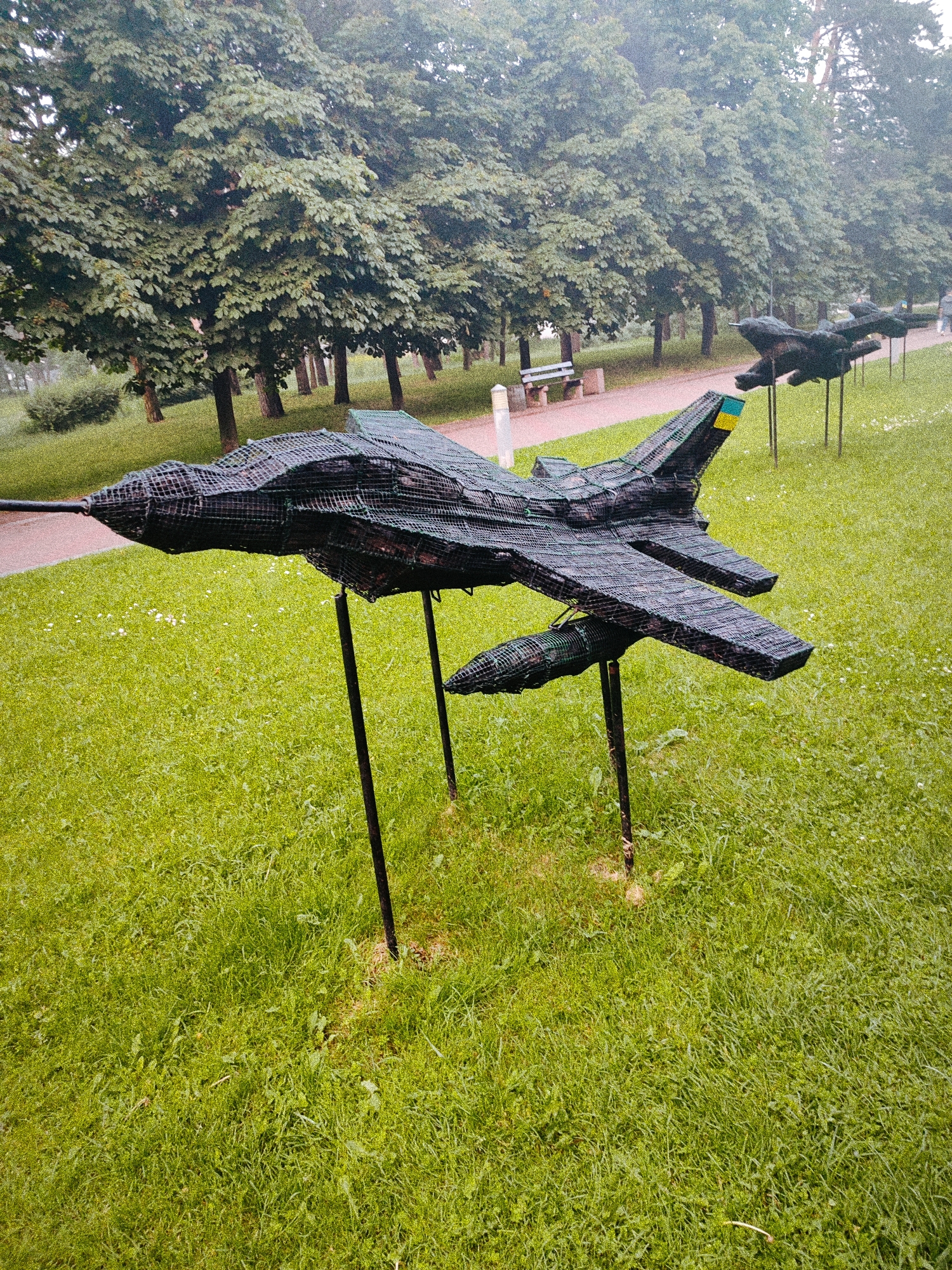 The image depicts an outdoor scene in a park in Kyiv, Ukraine. The foreground and midground consist of a vibrant green grassy area, interspersed with small white wildflowers. Positioned on the grass are three dark-colored sculptures resembling fighter jets, constructed from a mesh or netting material over a frame. The closest sculpture is prominently displayed on multiple dark support poles driven into the ground. Its tail fin features a vertical stripe with blue above yellow, indicative of the Ukrainian national flag. Two additional, similar jet sculptures are visible receding into the background along a parallel line. A light-colored paved pathway runs through the park, parallel to the jet sculptures and a dense line of large, mature deciduous trees with lush green foliage. A light-colored park bench and a waste receptacle are situated beside the pathway in the midground. An indistinct human figure is visible walking on the pathway in the distant background. The overall lighting suggests an overcast day.
