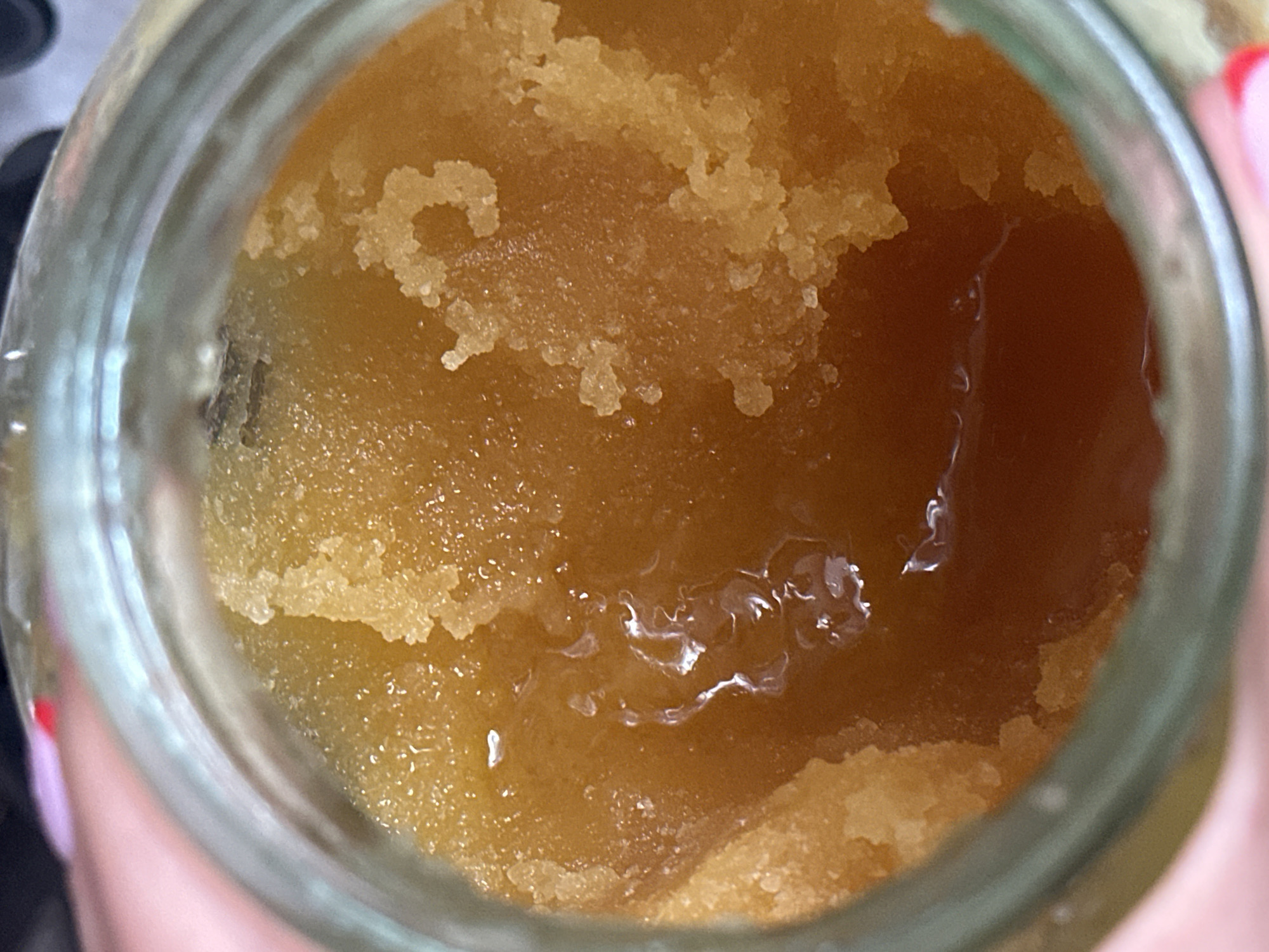 A close-up, overhead shot shows the inside of a glass jar filled with honey. The honey is a translucent amber color and has a granular texture with some liquid pooling on the surface. The jar rim is visible at the bottom and left edges of the image. The background is blurred and appears to be a person's hand with painted fingernails.