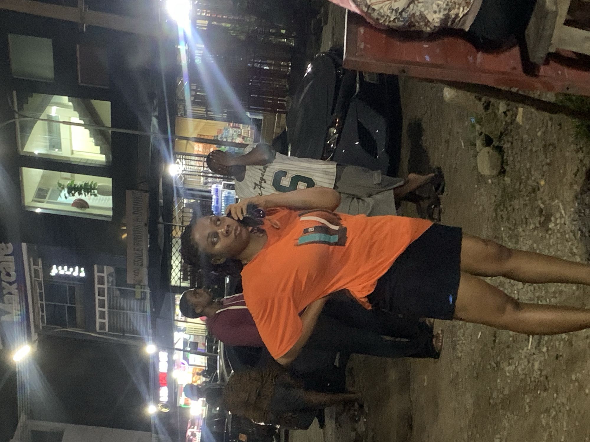 The image captures an outdoor scene at night in Galadimawa Village, Nigeria. A woman in an orange t-shirt and black skirt is the central figure, holding a phone to her ear with her right hand while resting her left hand on her hip. She appears to be engaged in a conversation. Behind her, a man in a white and green striped shirt with the number "6" on it stands with his hand over his face. To the left, a man in a burgundy hoodie is visible, and further left, another person in a patterned shirt and shorts is standing near a dark-colored car. The environment features a building with illuminated windows and signage that reads "Maxcare". Several cars are present, and the ground is uneven with gravel and debris. Streetlights cast a bright, somewhat hazy illumination across the scene, creating lens flares. A red structure, possibly a signpost or part of a stall, is visible on the right side of the frame.