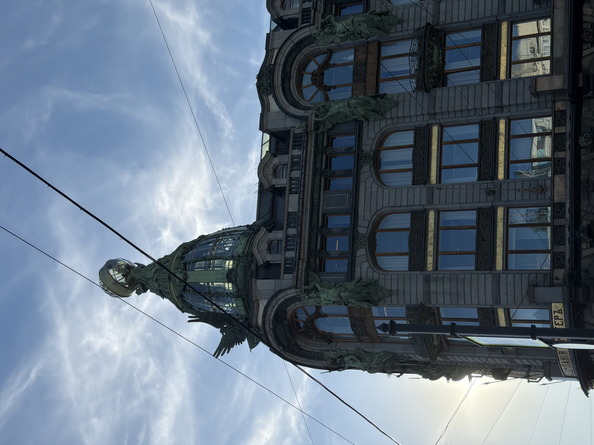 The image shows the Singer House, a historic building located at Nevsky Prospekt 20 in Saint Petersburg, Russia. The building features an iconic glass dome topped with a globe and an eagle sculpture. The facade is adorned with intricate architectural details, including statues of allegorical figures and ornate window frames. The sky is partly cloudy, with sunlight filtering through the clouds. Electrical wires are visible in the foreground, crossing the sky. The perspective is from below, looking up at the building. The photograph captures a clear day with good visibility.
