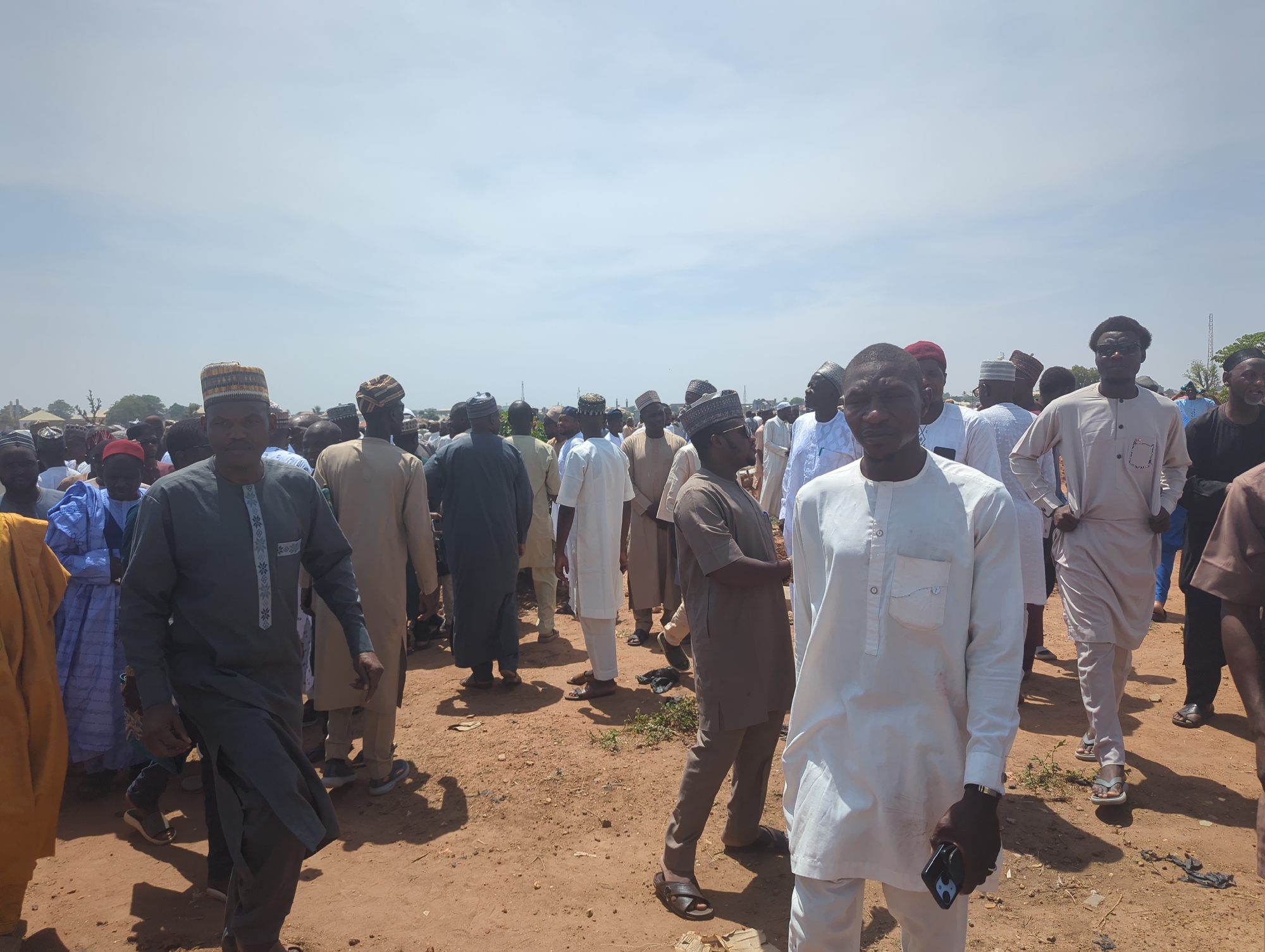 The image captures a large outdoor gathering of men in Sardauna, Nigeria, under a bright, sunny sky with scattered clouds. The ground is a reddish-brown, dusty earth with some sparse patches of green vegetation and shadows indicating midday or early afternoon.
The subjects are numerous men, predominantly Black/African, dressed in traditional attire. Many wear long, flowing garments like kaftans and robes in various colors, including white, grey, brown, light blue, and yellow. A significant number of men are also wearing kufis (traditional caps) on their heads.
In the foreground, several individuals are clearly visible. On the right, a man in a white kaftan with a matching cap stands looking towards the viewer, holding a black smartphone in his left hand. Next to him, a man in a brown outfit and cap stands with his arms crossed. On the left, another man in a dark grey kaftan and a striped kufi is walking towards the foreground. Most faces are neutral, with some individuals looking directly at the camera.
The crowd extends deep into the background, suggesting a substantial gathering. In the distant background, faint outlines of trees and possibly some structures can be seen. The overall scene conveys a sense of a community event or social assembly. There is no visible text in the image.