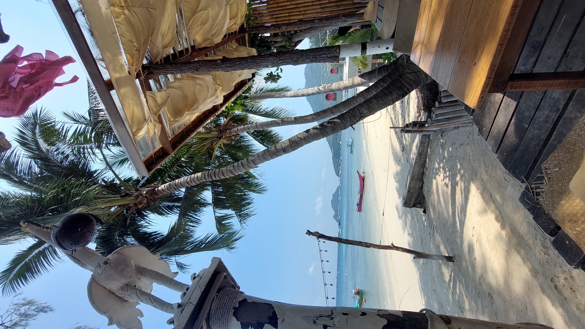 The image captures a coastal scene in Koh Tao, Thailand, taken from an elevated position within a beachside structure. Visible are palm trees and their fronds, reaching towards a clear, blue sky. A white painted sculpture of a bird stands in the foreground. Parts of the beachside structure include a bamboo-lined awning and a wooden table and deck. The beach itself is white sand, stretching towards the sea, where several boats are visible. Further out, the outline of distant islands is discernible. A string of decorative lights stands on the beach.